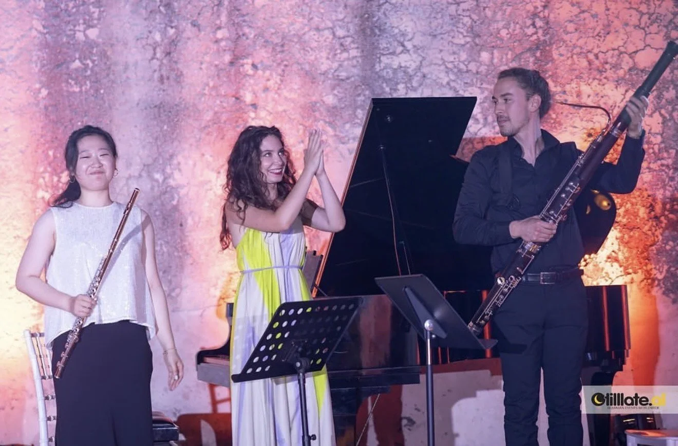 Three musicians on stage, two women and one man, with a grand piano and music stands, in front of a textured mauve curtain background.