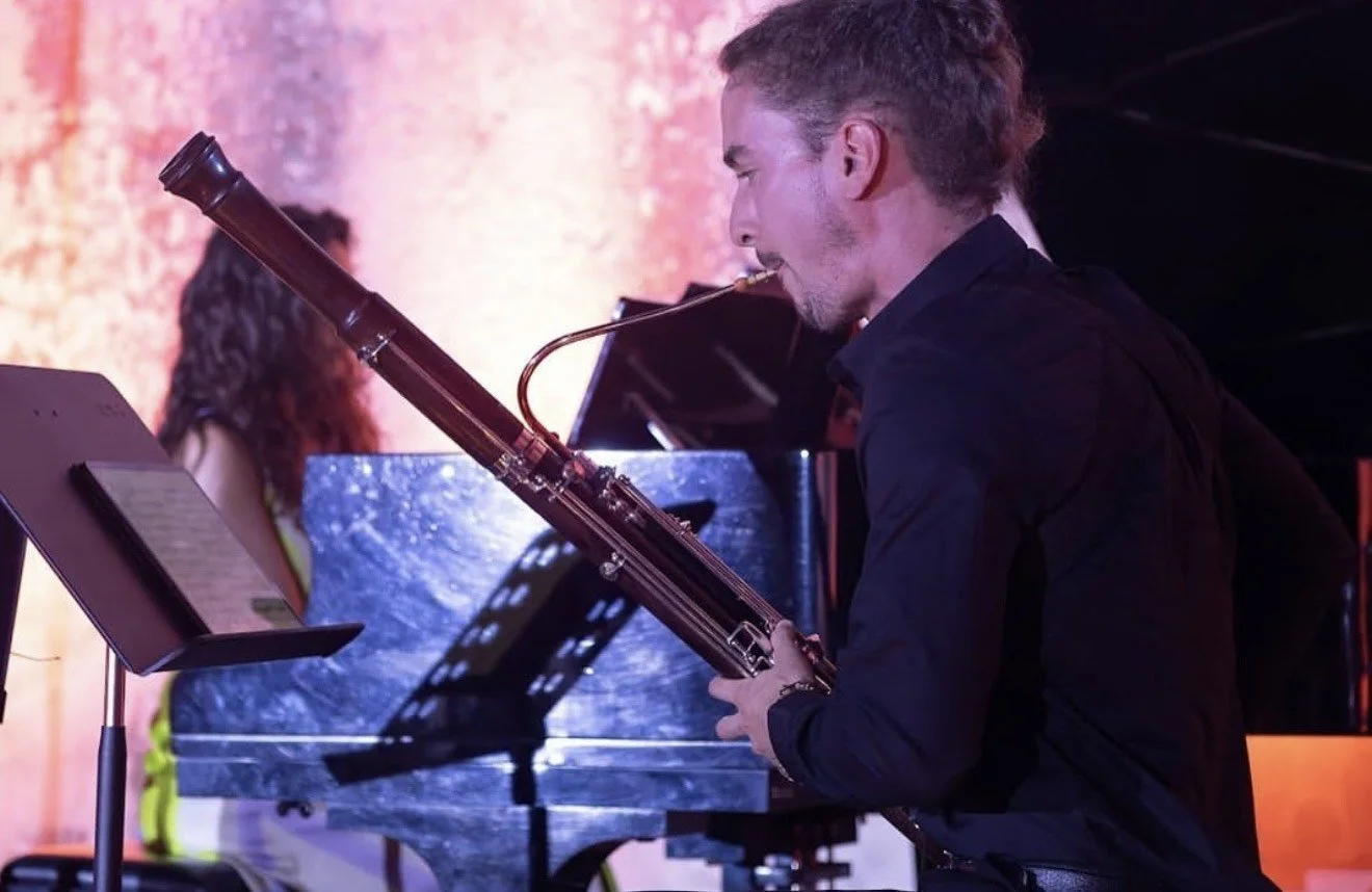 A man playing a bassoon during a musical performance with a pianist in the background.