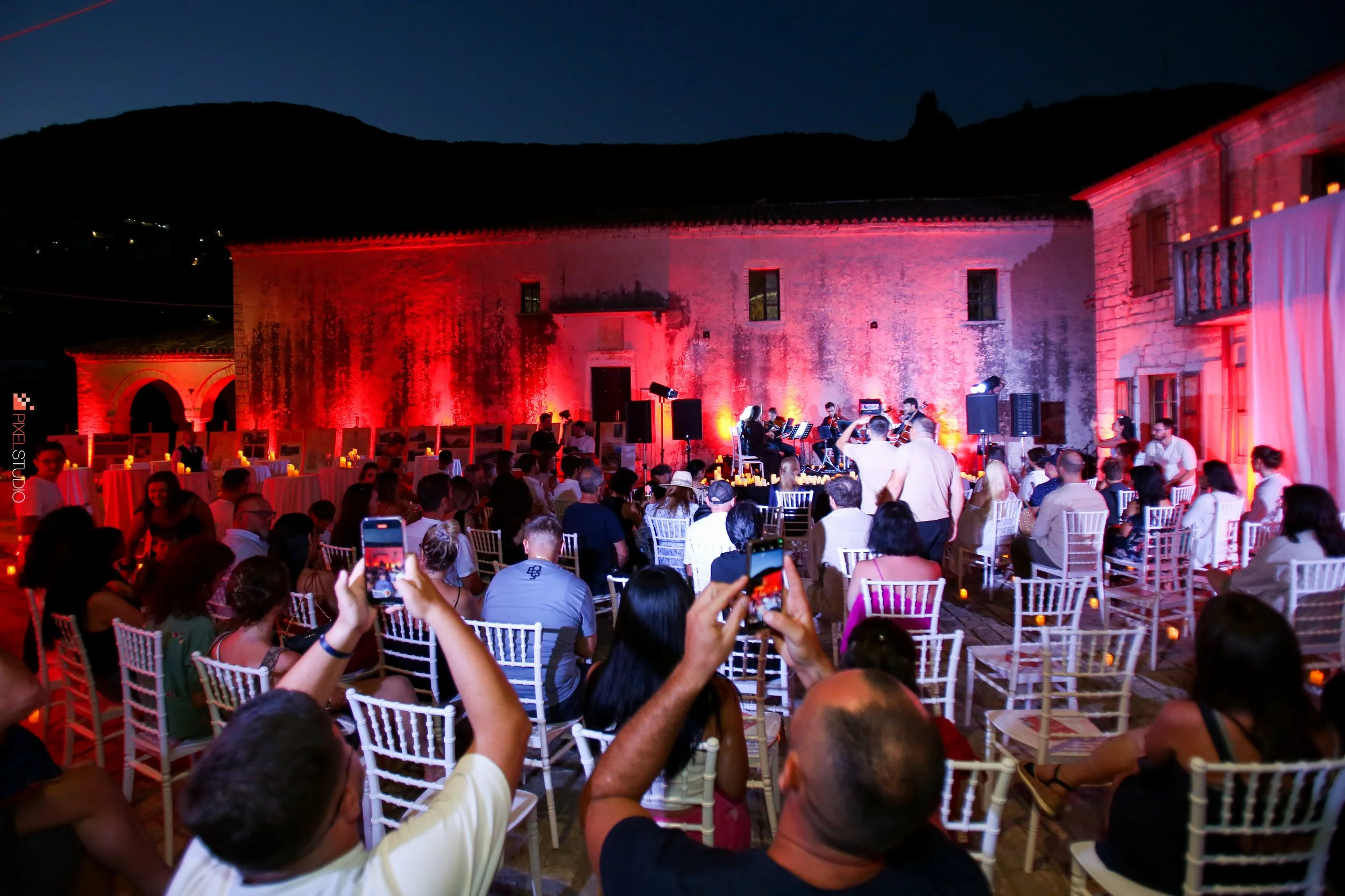 An outdoor evening event with a large crowd seated on white chairs, watching a musical performance on a small stage illuminated with colored lights. The background features a historic stone building with a red hue cast on its walls, and the dark blue sky indicates it is nighttime.