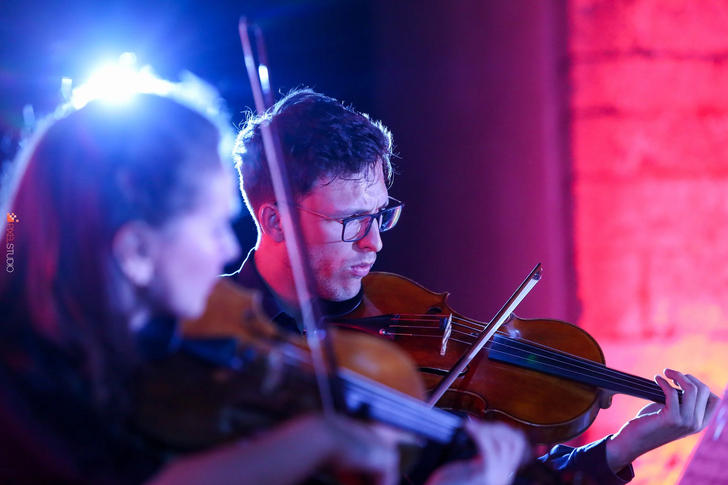 Two musicians playing violins in a dim, colorful stage setting with blue and pink lighting.