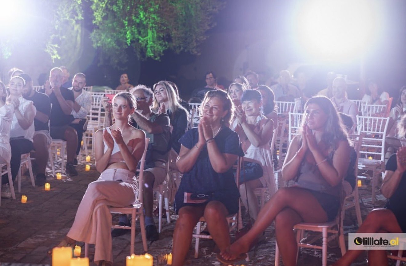 An outdoor evening event with an audience seated on white chairs, illuminated by small candles on the ground. The audience, consisting of men and women, is clapping and appears to be enjoying a performance, with some smiling and others focused. Bright lights shine from behind the audience, creating a festive atmosphere.