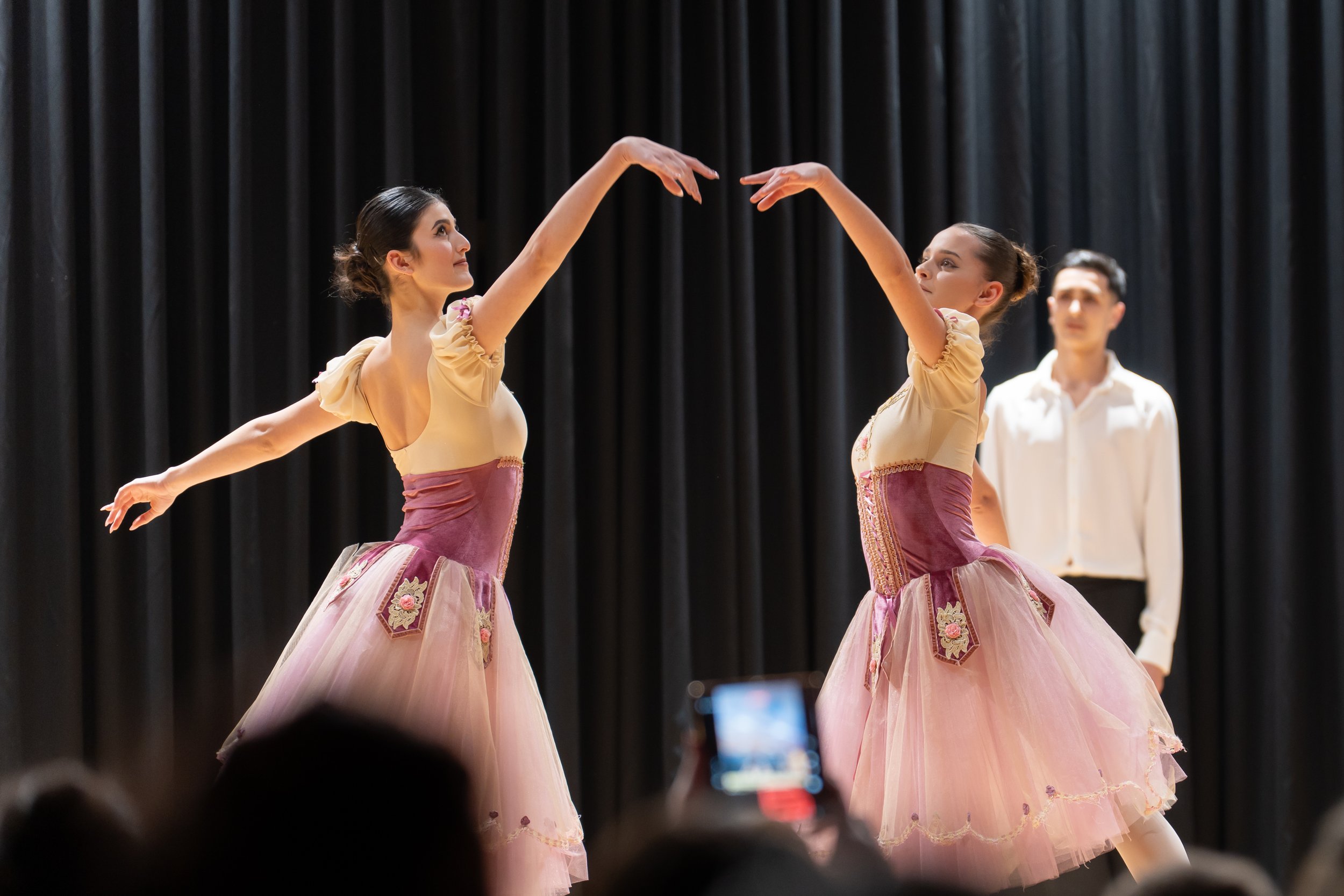 Two female ballet dancers in pink costumes performing on stage with black curtains in the background, while a man in a white shirt stands behind them.