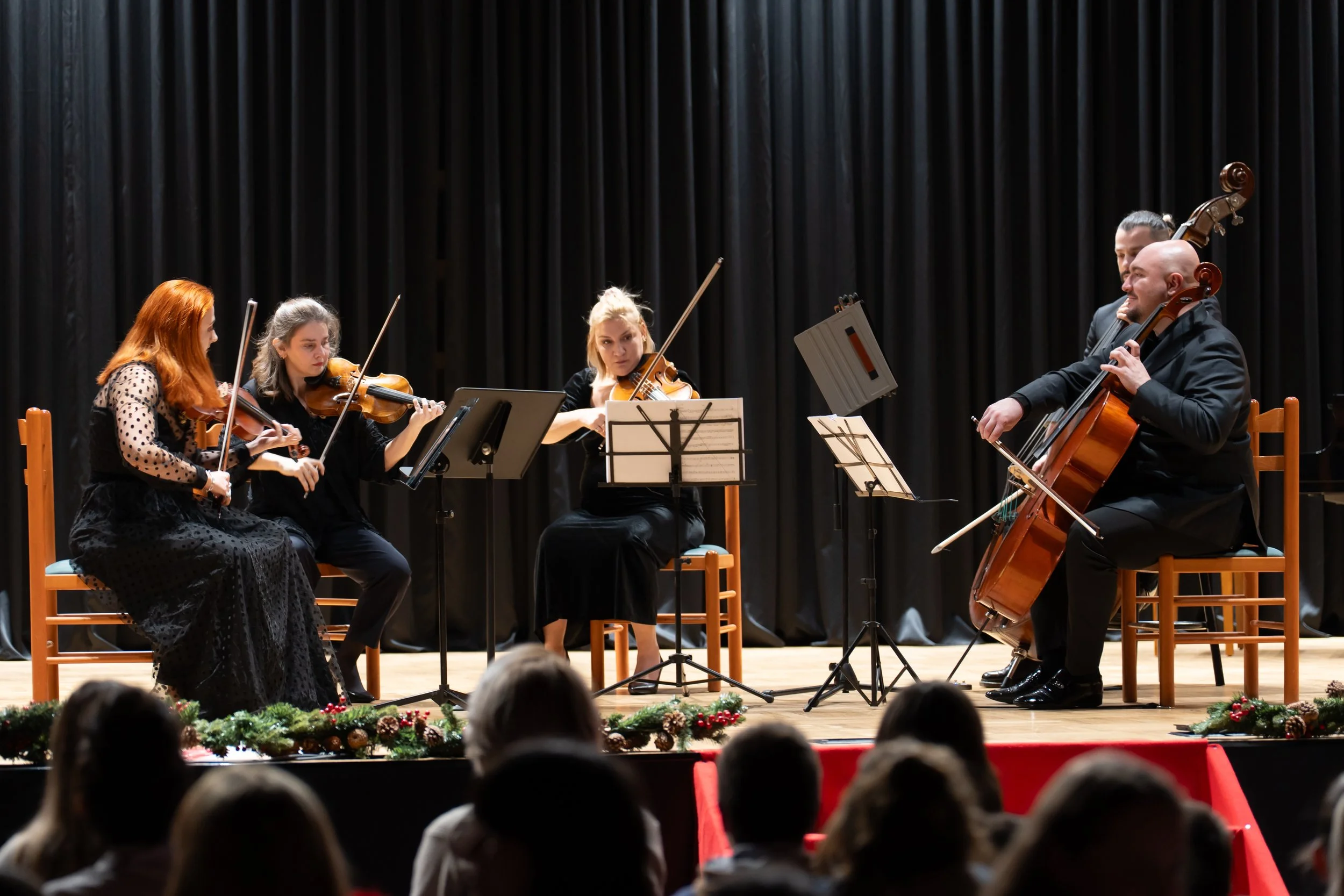 A group of five musicians performing on stage with string instruments, including a violin and double bass, during a concert with a black curtain backdrop and festive decorations at the front of the stage.