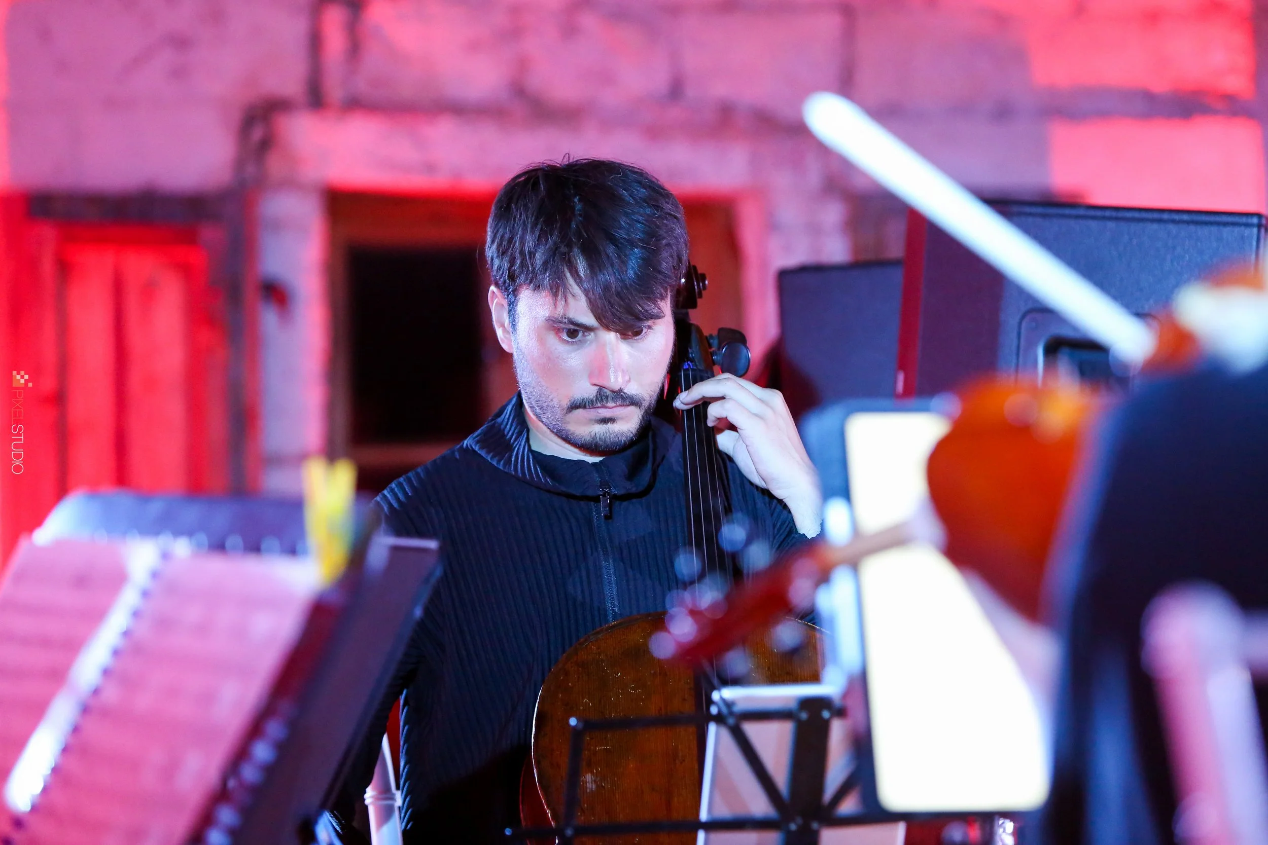A man with dark hair and a beard playing the cello, focused on his instrument, with a red-lit background.