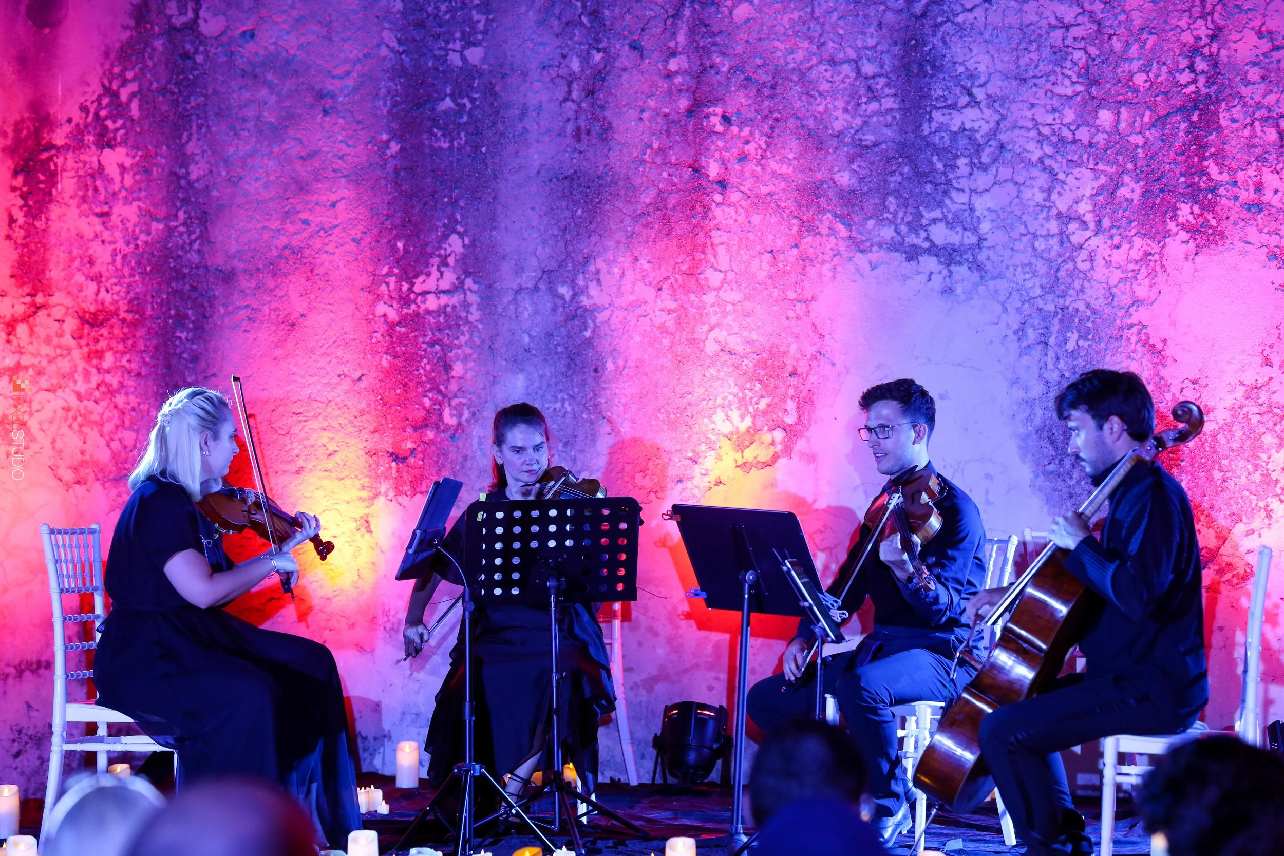 A group of five musicians playing string instruments on a stage with a colorful textured background lit in pink and purple hues, candles on the floor.