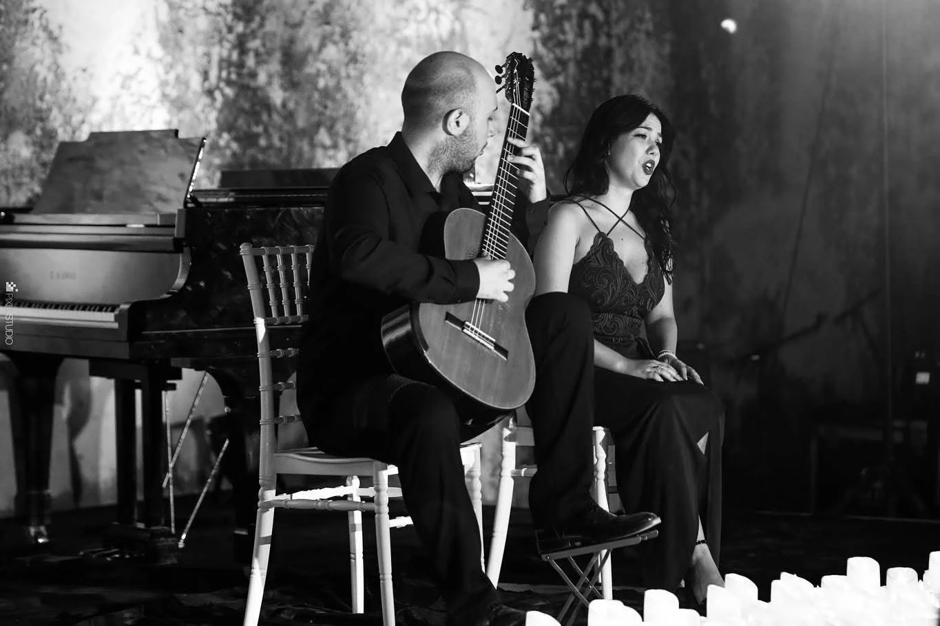Black and white photo of a man playing guitar and a woman singing on stage, with piano in background, candles on floor in foreground.