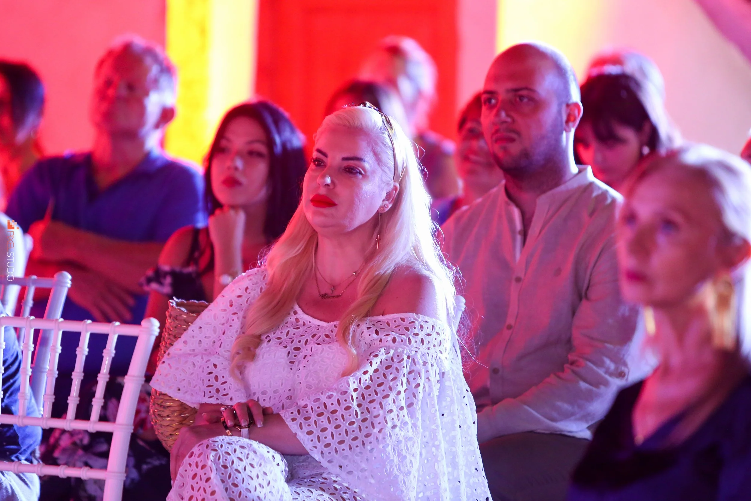 A group of people sitting in a brightly colored room, watching something attentively, with a woman in the foreground wearing a white, eyelet dress and red lipstick.