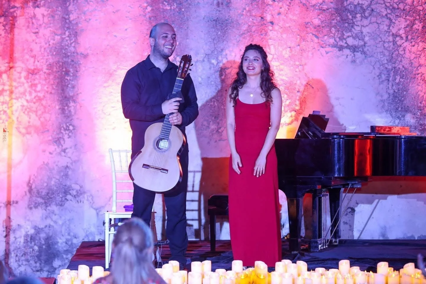 A man with a guitar and a woman in a red dress standing on stage with a grand piano, surrounded by candles, against a textured wall illuminated with pink and purple lighting.