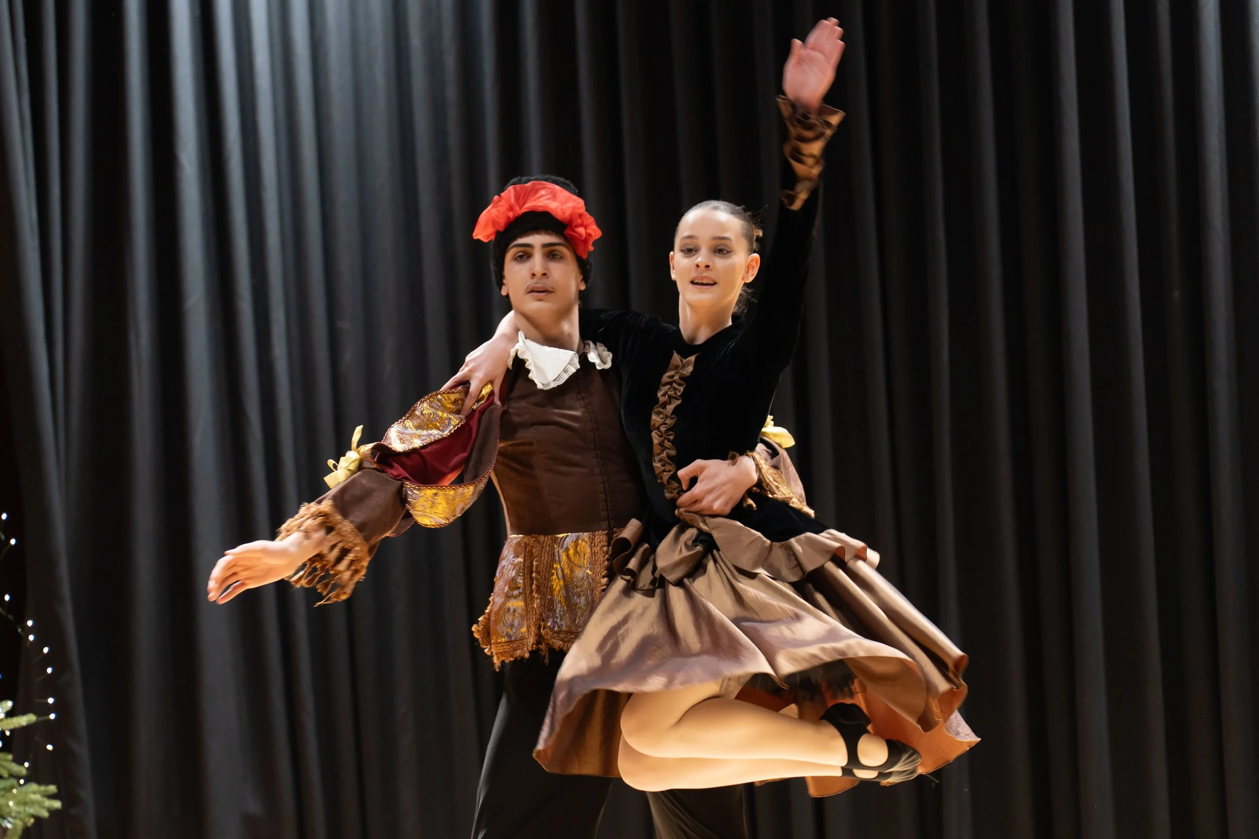 Two ballet dancers performing on stage, dressed in elaborate period costumes against a dark curtain background.