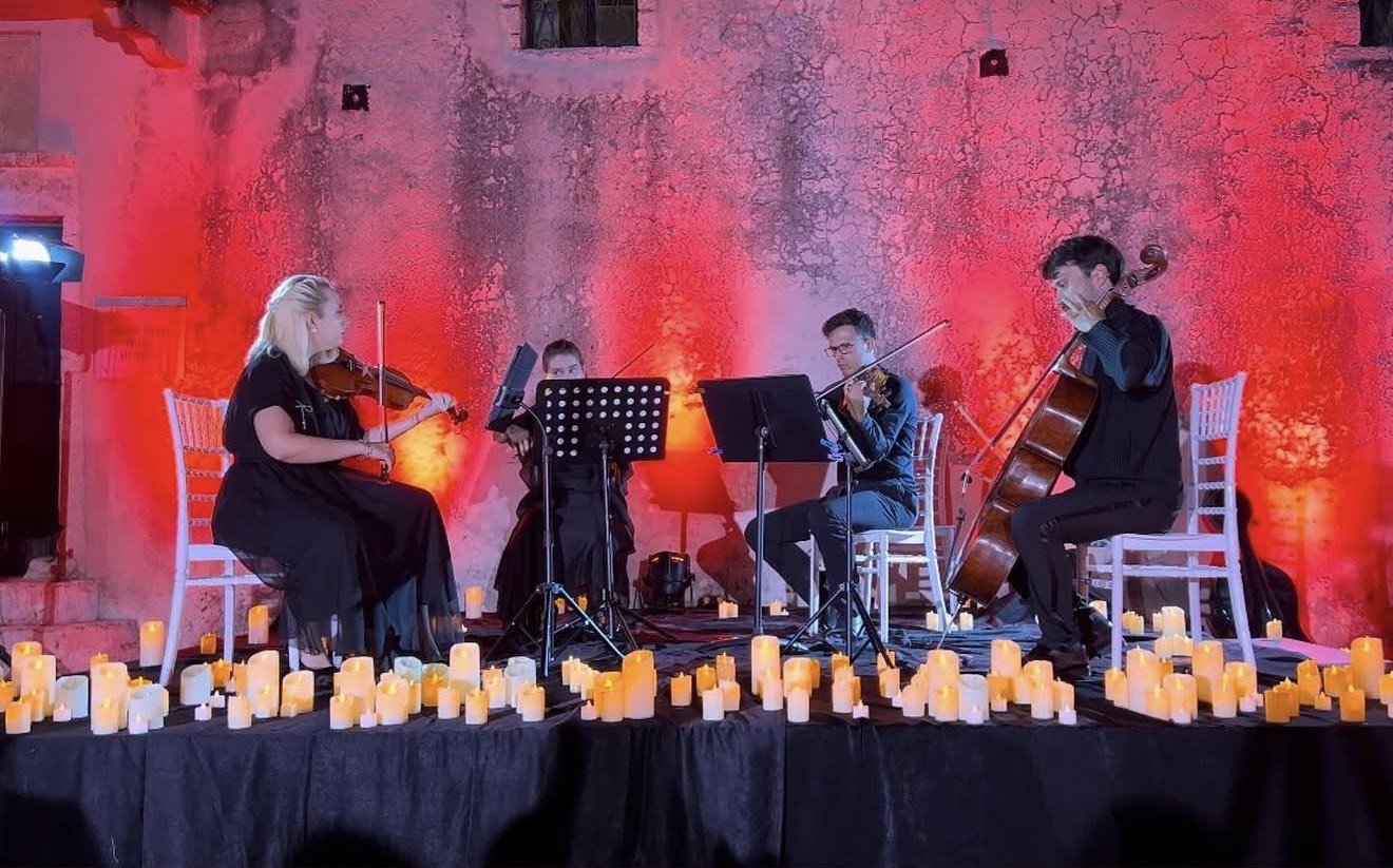 A string quartet performing on stage with candles in front, against a textured red illuminated wall, with two women and two men playing violin, viola, and cello.