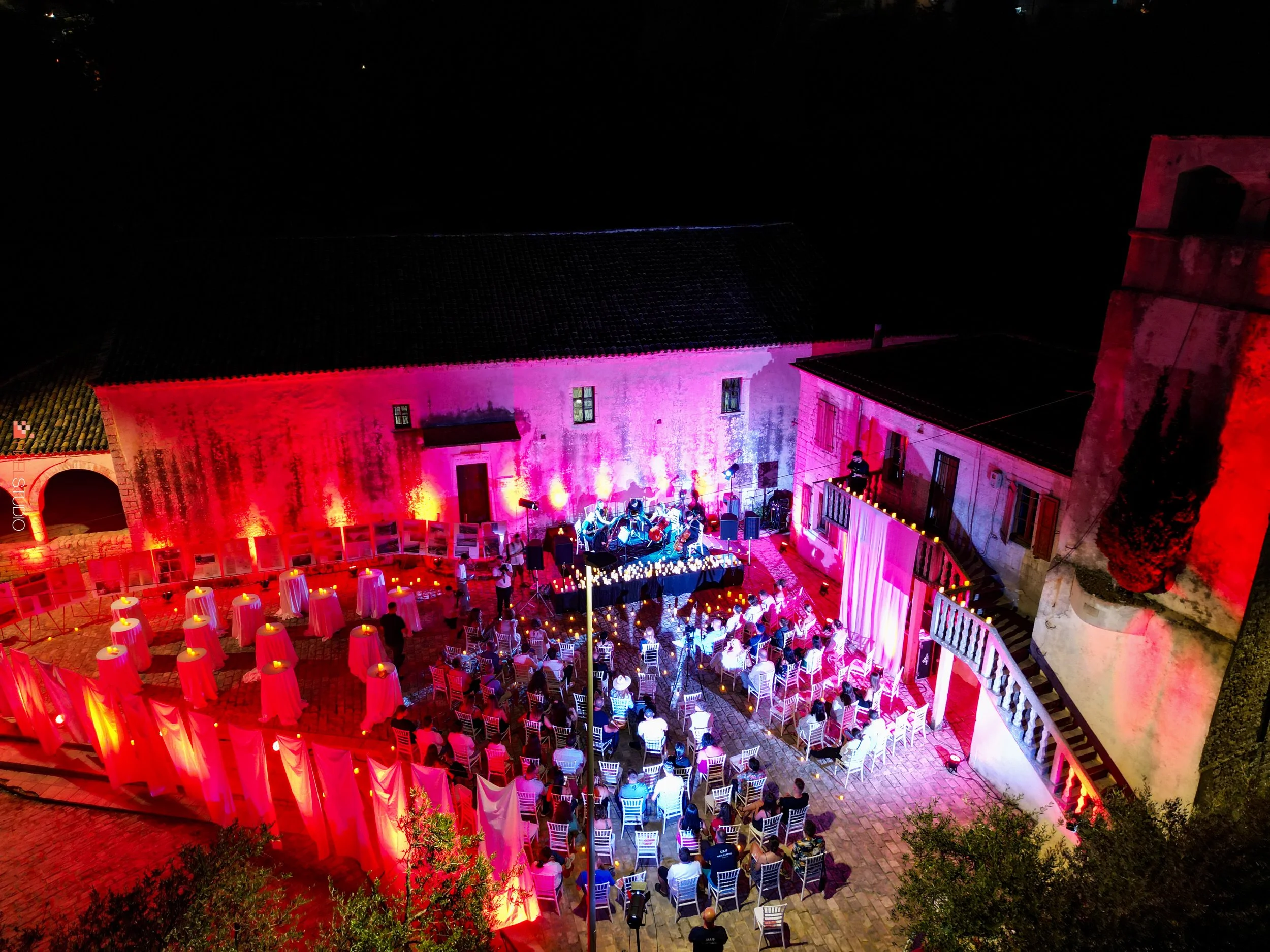 A nighttime outdoor event with people seated on white chairs, illuminated by pink and purple lighting on the walls of old stone buildings, with some standing and mingling, in a courtyard with a stage and musical performers.