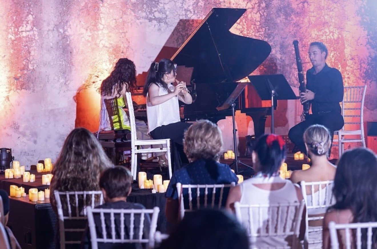 Young girl playing the recorder, woman playing the clarinet, man playing the bassoon performing on stage at a concert surrounded by audience members with lit candles on tables.