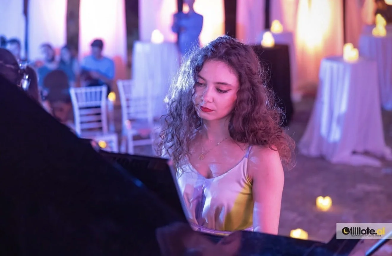 A young woman with curly hair in a satin dress playing piano at a dimly lit event or party with candles and guests in the background.