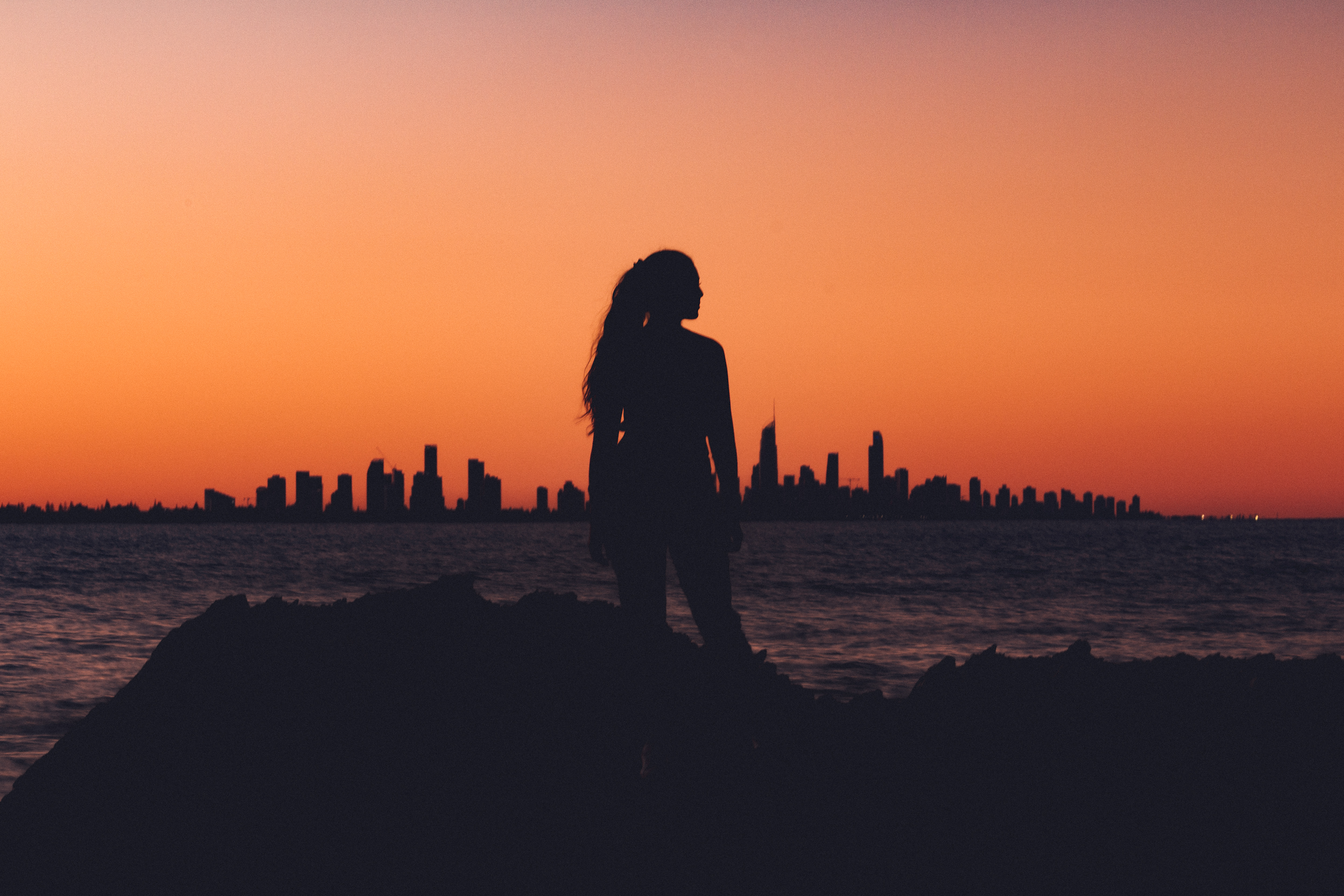 Silhouette of a woman standing on rocks near water at sunset, with a city skyline in the distance.