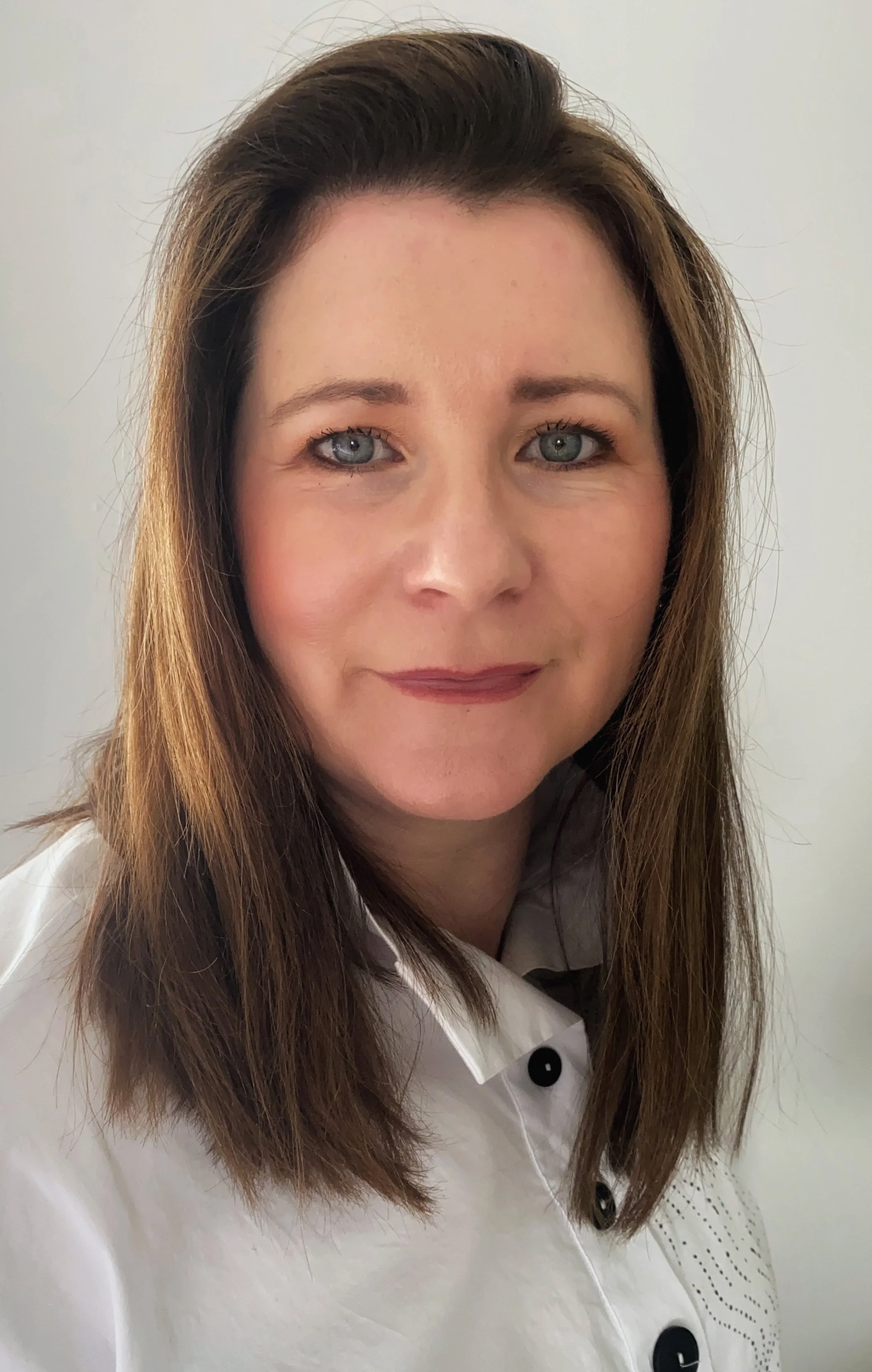 A woman with long brown hair, blue eyes, and light makeup wearing a white shirt with black buttons and black embroidered details, standing against a plain light background.