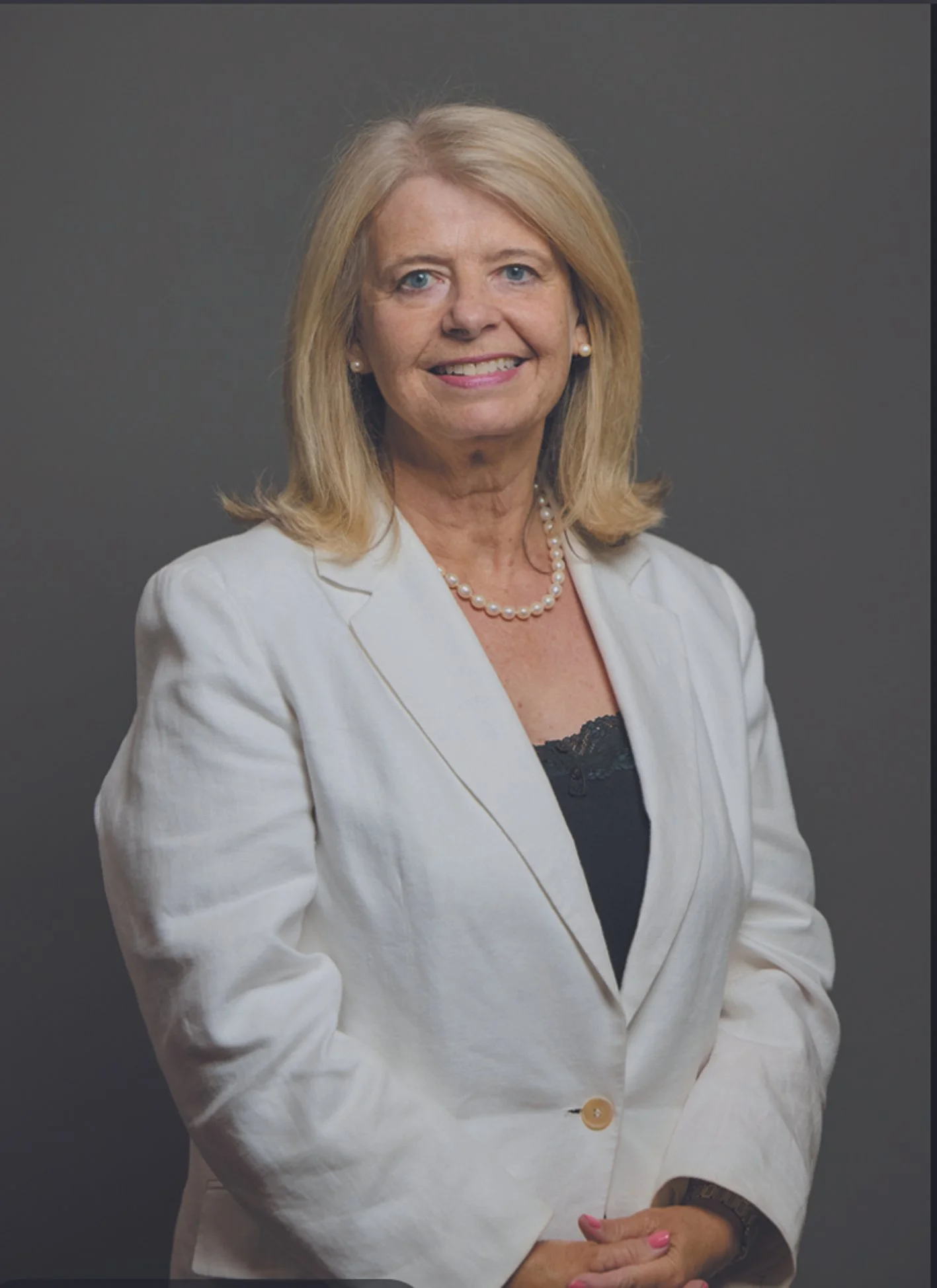 Portrait of a smiling woman with blonde hair wearing a white blazer, pearl necklace, and earrings, standing against a gray background.