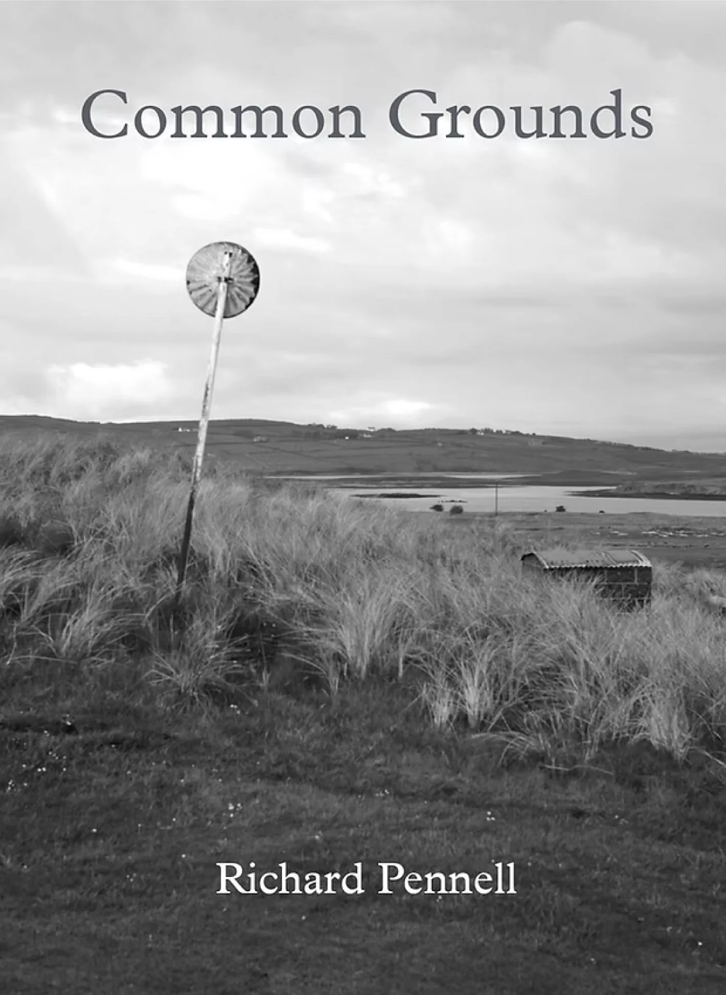 Book cover titled 'Common Grounds' by Richard Pennell, showing a rural landscape with tall grass, a tilted windmill, a small shed, and a lake or pond in the background in black and white.