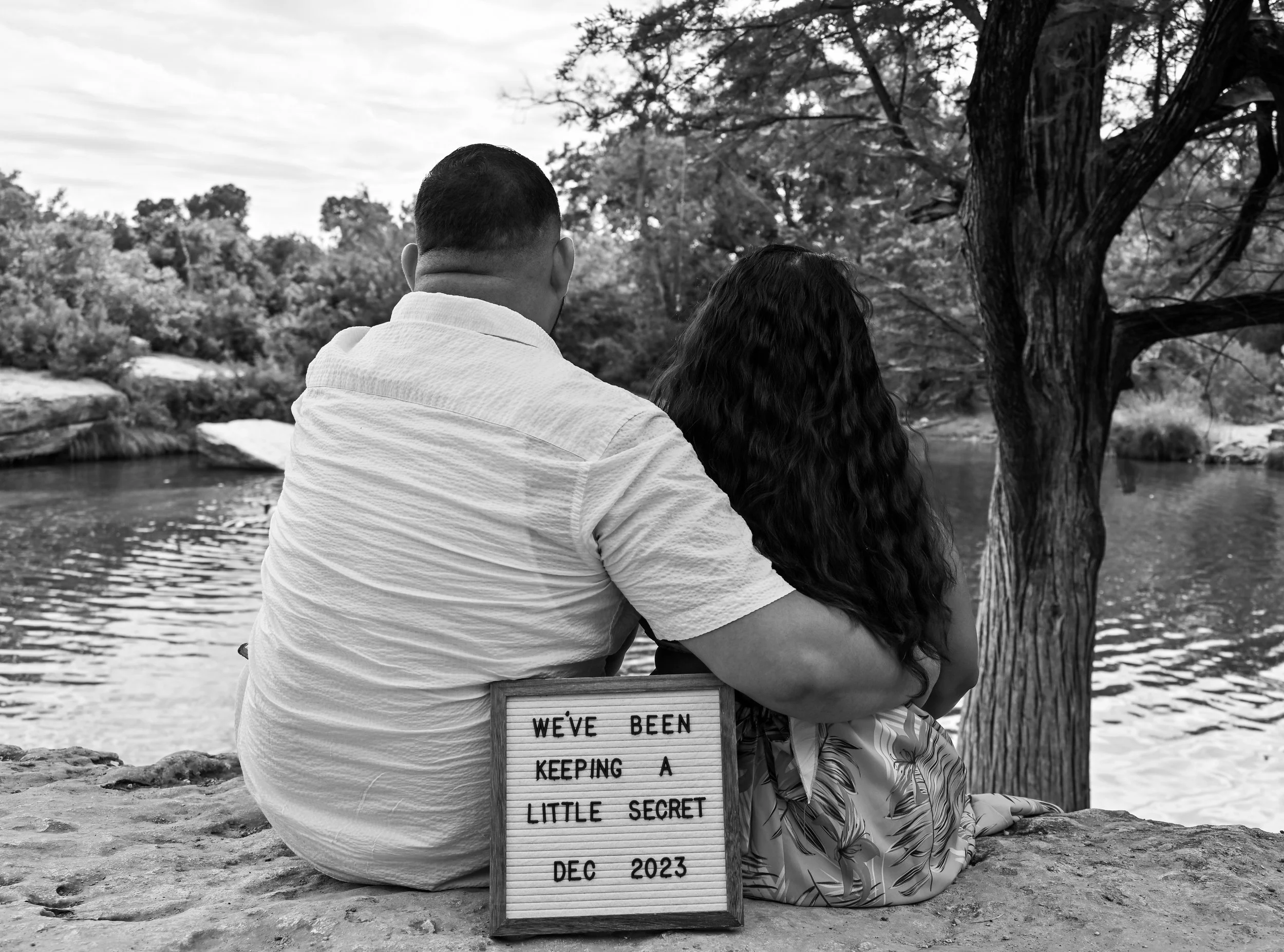 Expecting parents sitting beside a river revealing their pregnancy announcement sign during a maternity photography session in Austin, Texas by Caerus Multimedia.