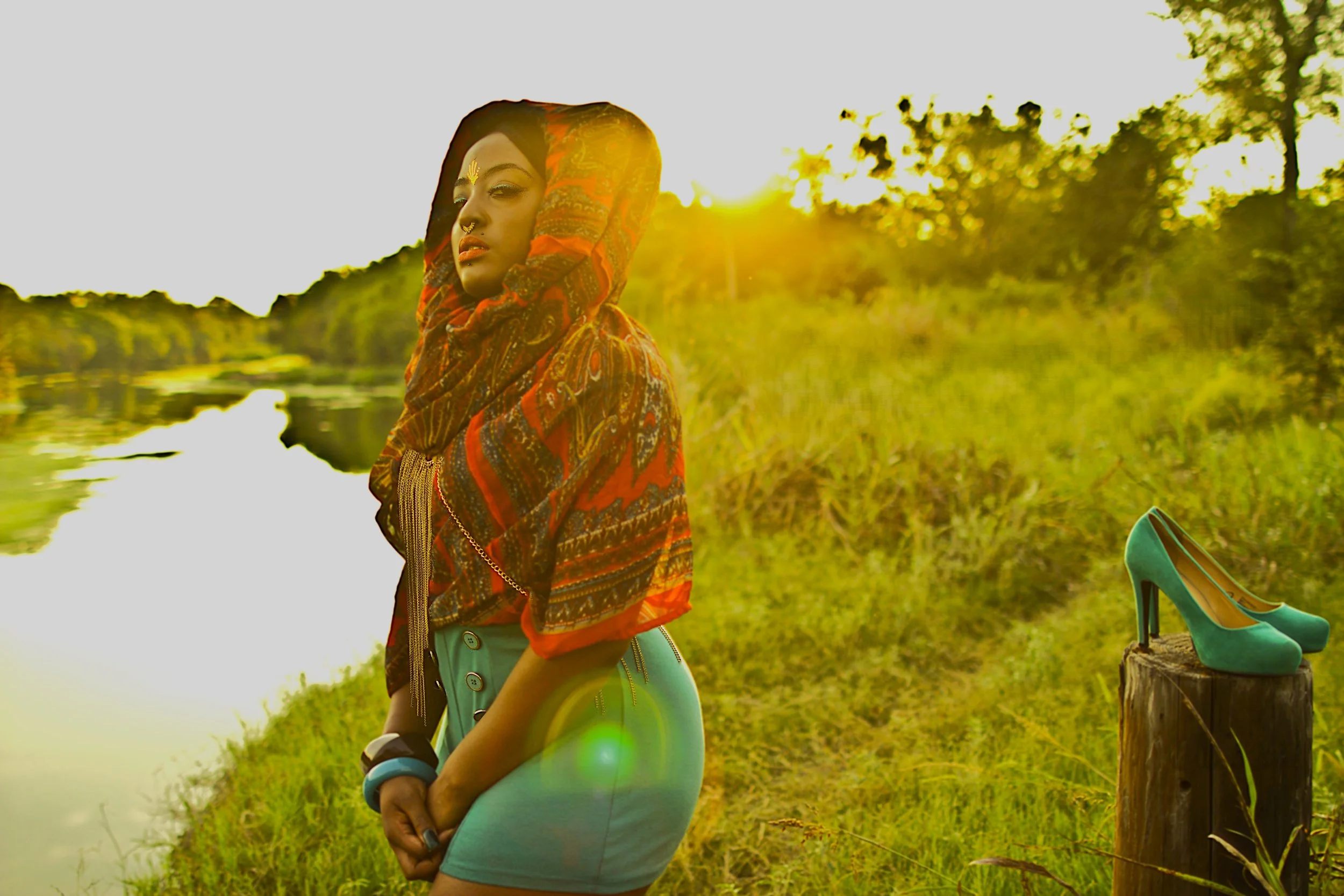 Standing at the edge of the water as the sun sets behind her, the Queen of the Golden Sun reflects in a moment of quiet strength and transformation. The warm golden light and natural landscape frame her presence, while the shoes set aside symbolize g