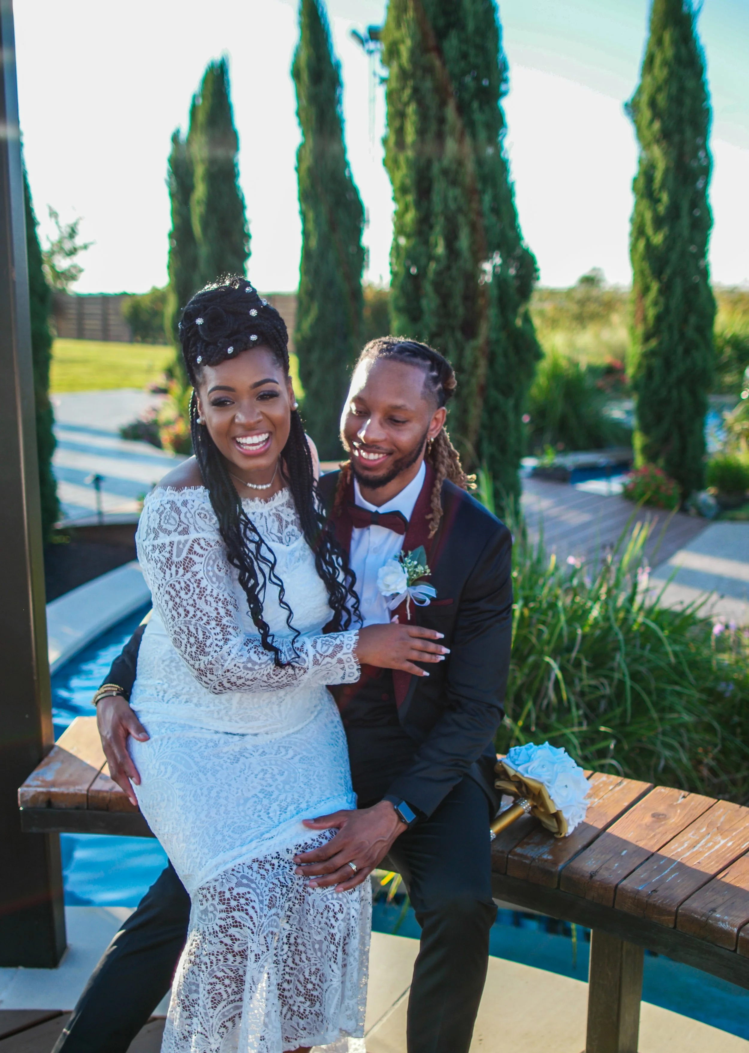 The bride and groom share a joyful embrace beside a garden walkway surrounded by tall cypress trees and warm sunlight. This portrait captures the romantic atmosphere and candid connection that Caerus Multimedia wedding photography is known for.
