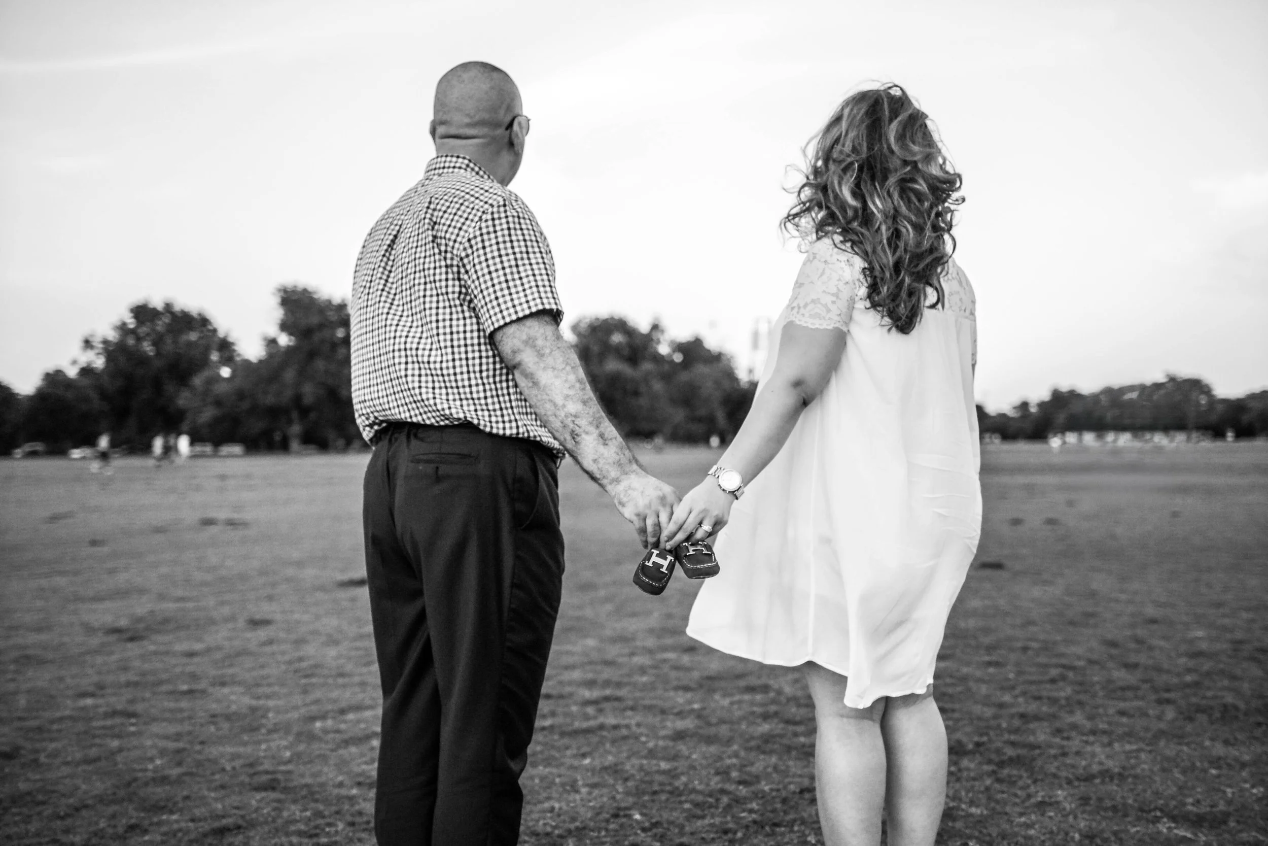 Expecting parents walking together across an open field holding baby shoes during a maternity photography session in Austin, Texas. A storytelling pregnancy portrait photographed by Caerus Multimedia.