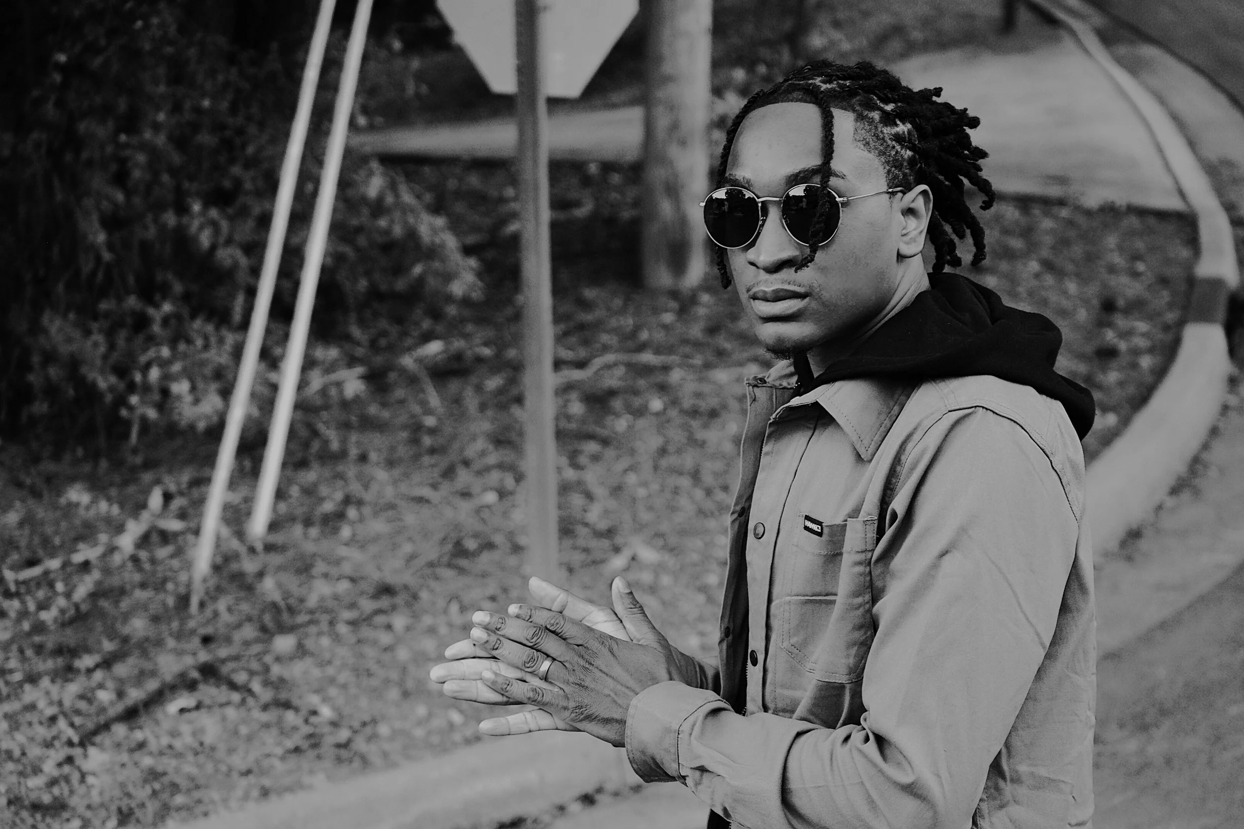 A dramatic black and white portrait featuring strong contrast and thoughtful composition. The subject’s stance and expression bring intensity to the image while the natural background adds depth and atmosphere.