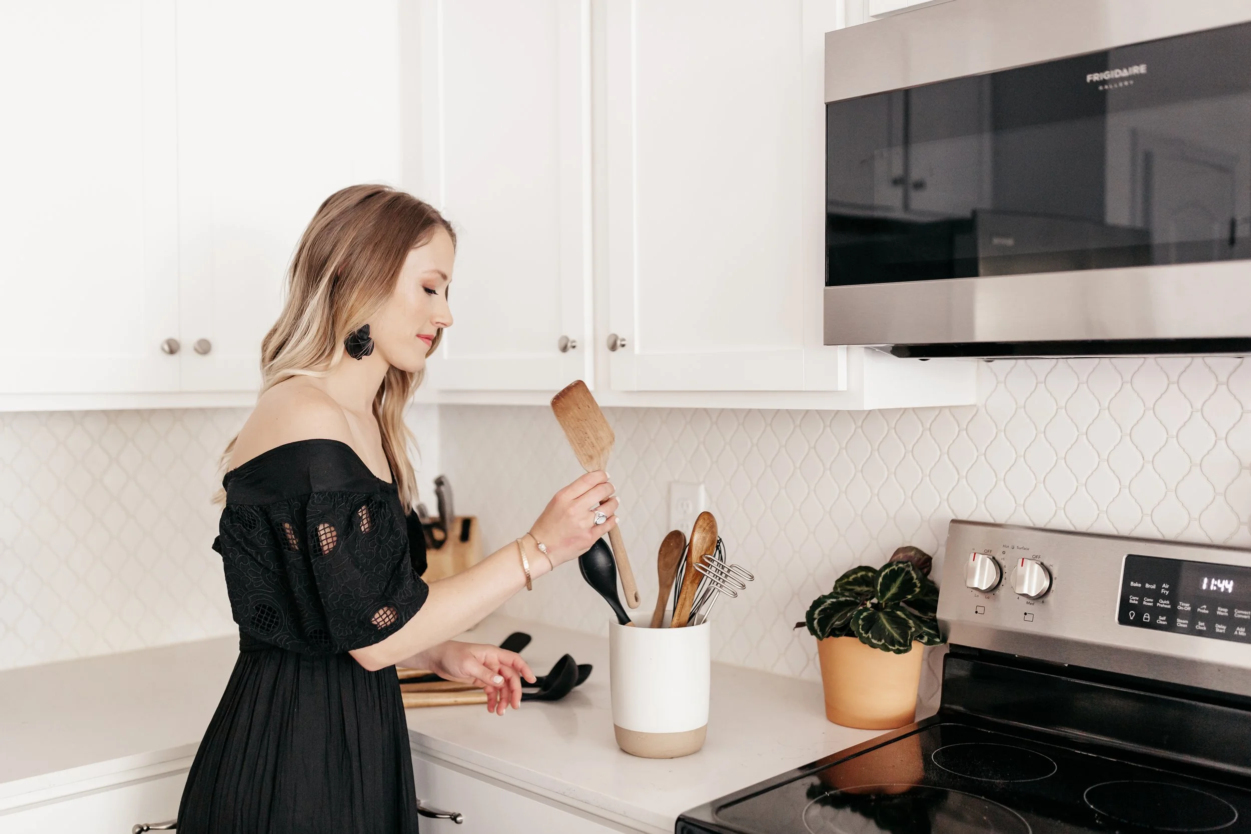 A woman with long hair wearing a black dress and large earrings is cooking in a white kitchen. She is holding a spatula and standing next to a vent hood over an electric stove.