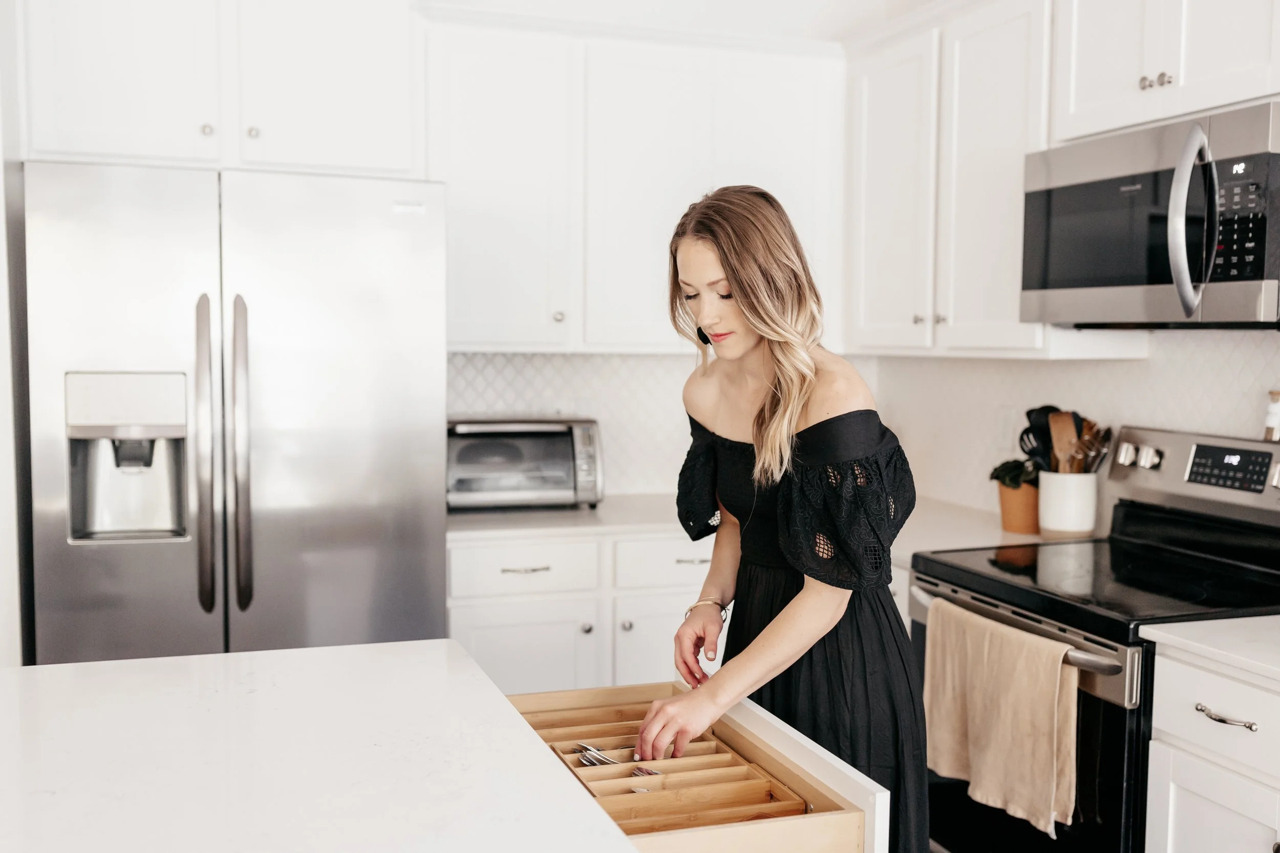 A woman with long, wavy, light brown hair wearing an off-the-shoulder black dress, standing in a modern white kitchen with stainless steel appliances, opening a wooden cutlery drawer.