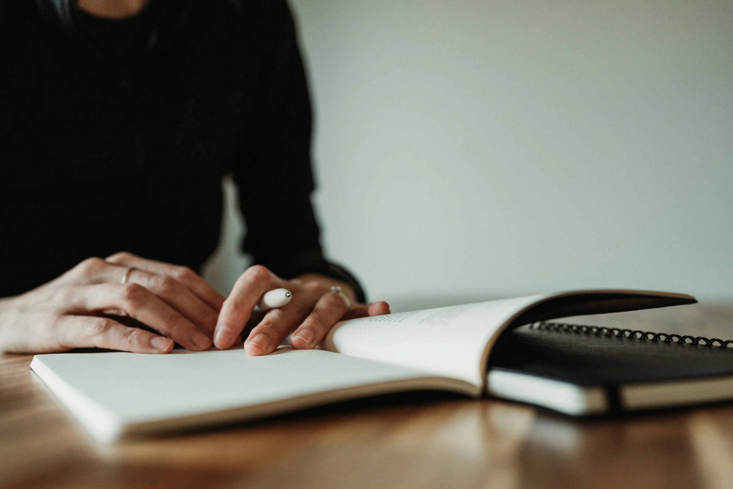 A person wearing a black shirt writing in a notebook with a white pen on a wooden table.