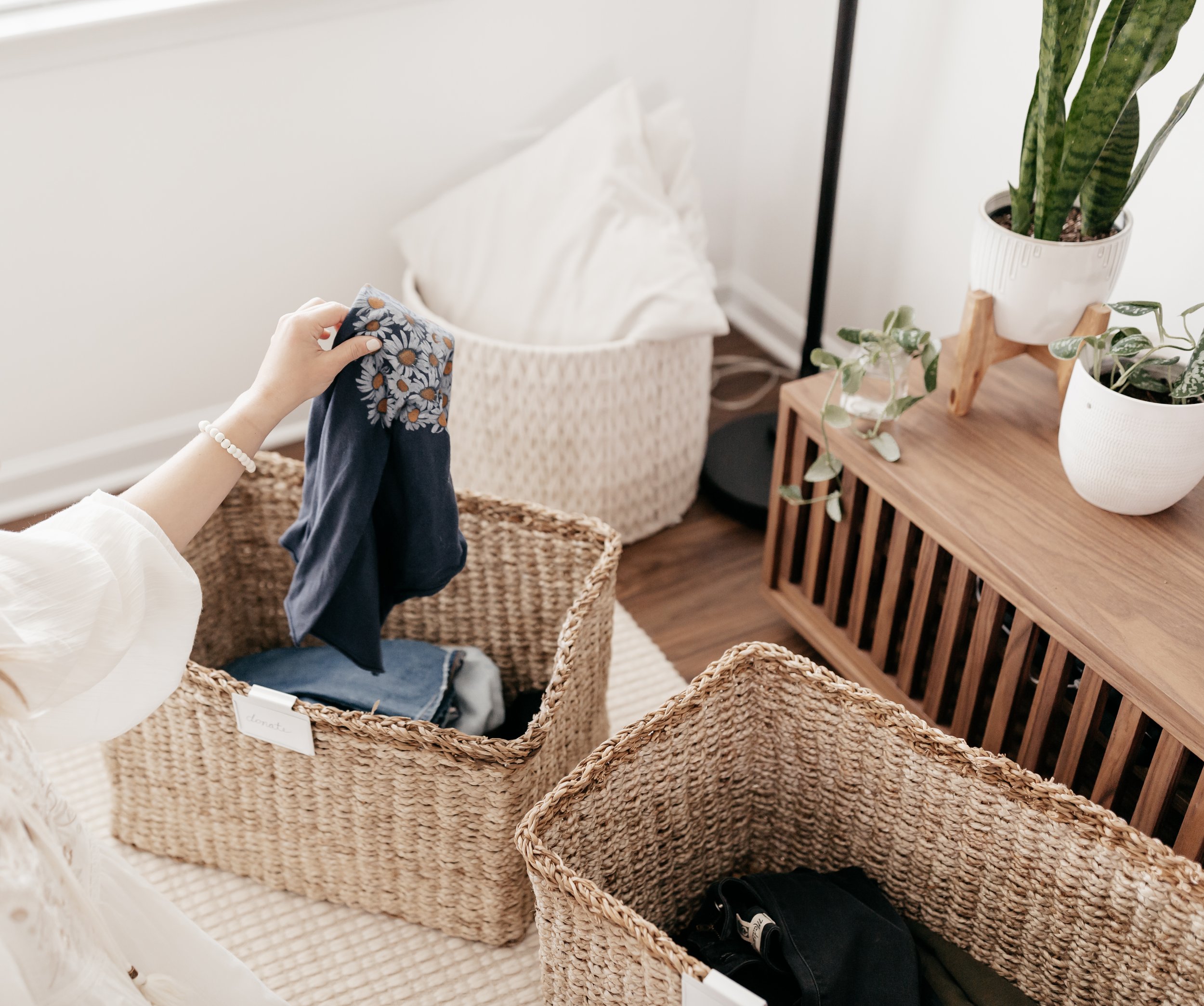 Person folding a dark blue cloth with floral pattern in a wicker basket inside a room with wooden furniture and potted plants.