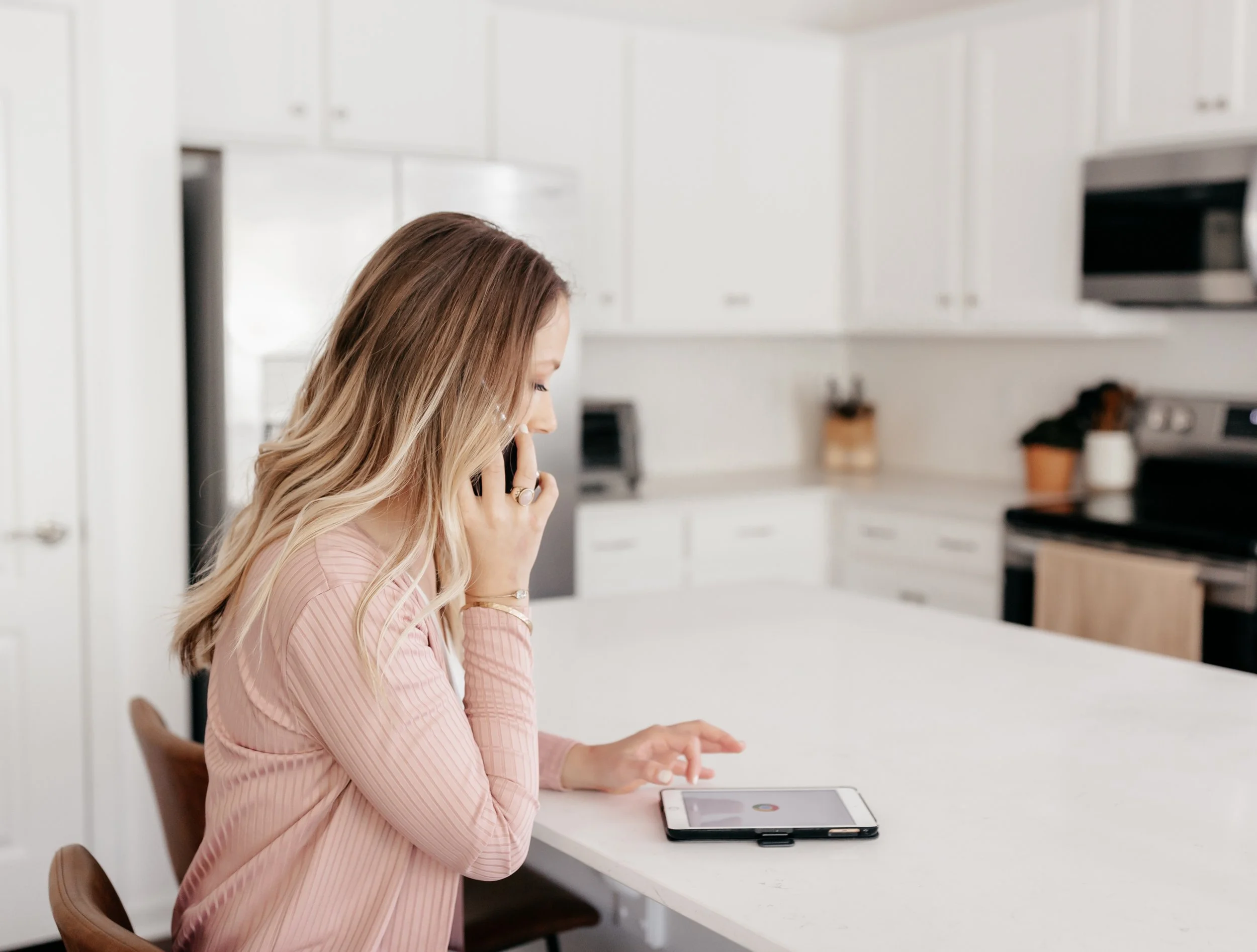 A woman with long blonde hair wearing a pink ribbed dress sitting at a white kitchen counter, talking on her mobile phone while using a tablet.