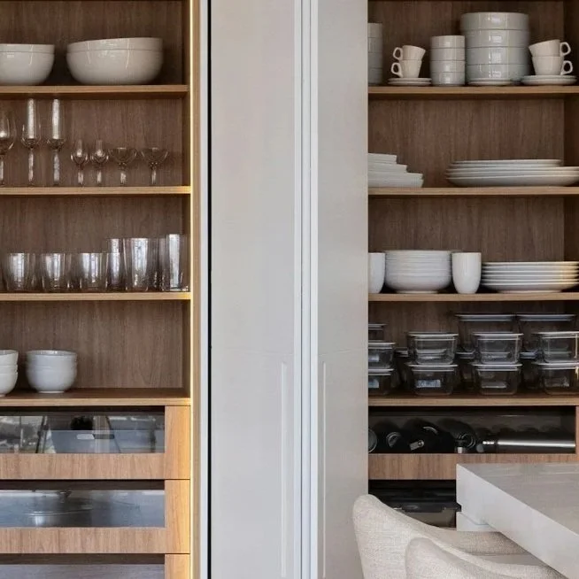 Kitchen cabinets with open shelves filled with white dishes, bowls, glasses, and clear containers, with a chair and part of a counter visible in the foreground.