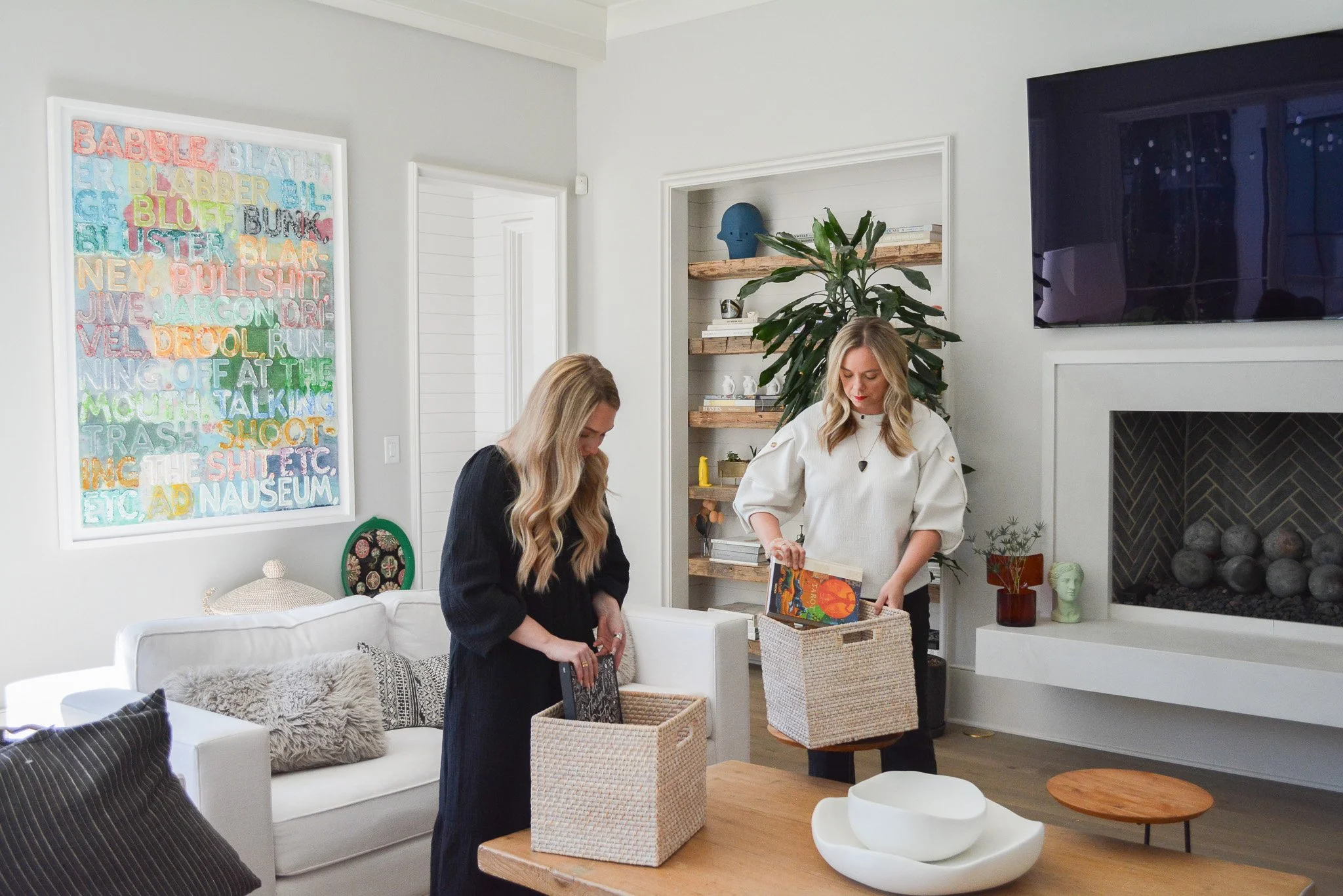 Two women organizing items in a living room with white walls, artwork, a large plant, fireplace, television, and decorative objects.