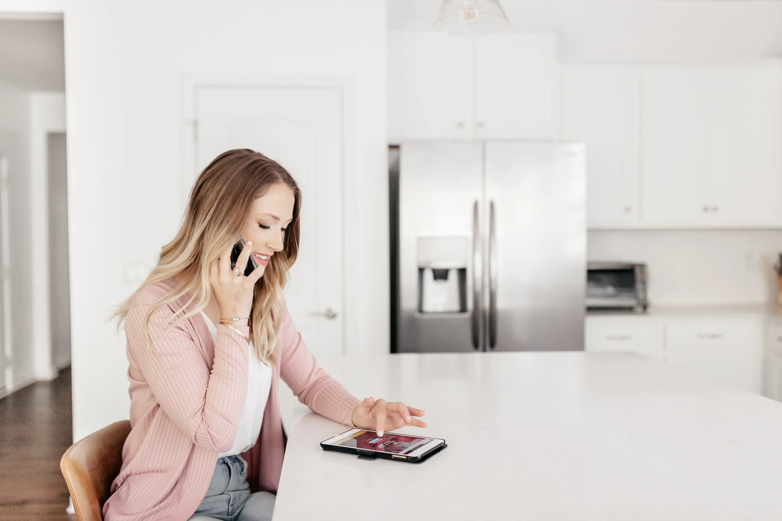 A woman sitting at a white kitchen countertop, talking on a cellphone and using a tablet device.