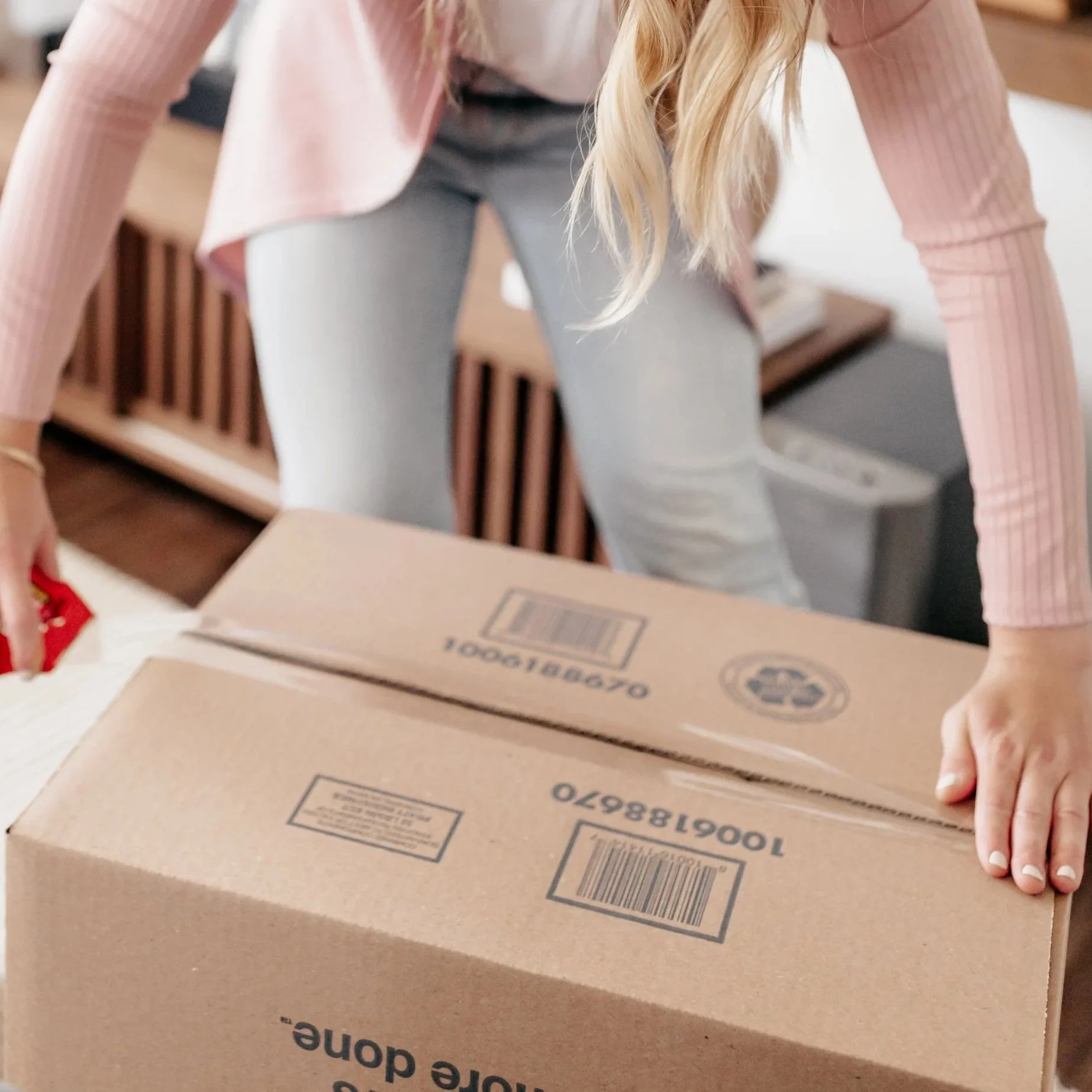 Person in a pink and white striped long-sleeve shirt and gray pants leaning over a cardboard box, with long blonde hair partially covering their face.