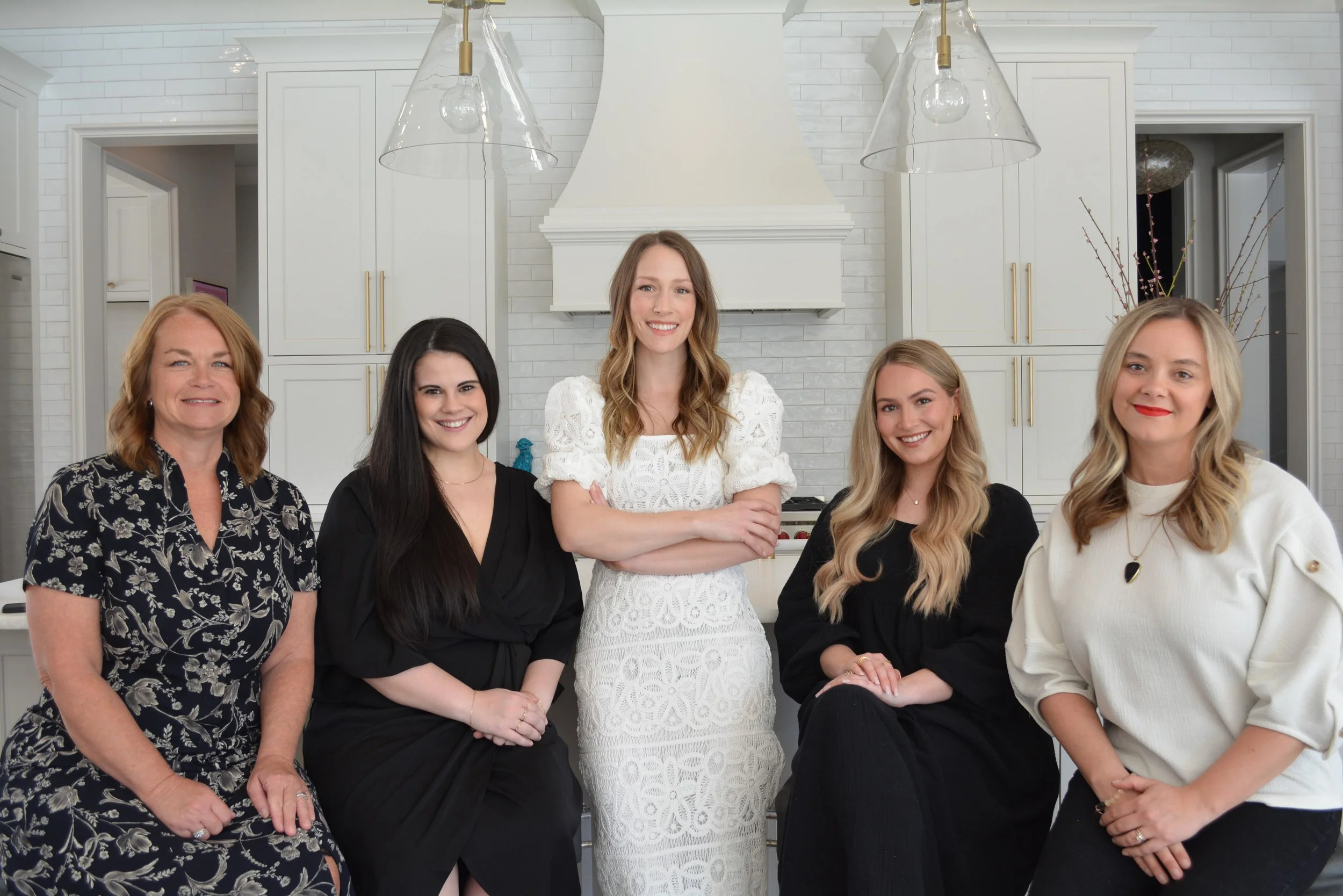 Group of six women smiling in a modern kitchen with white cabinets and a white hood vent.