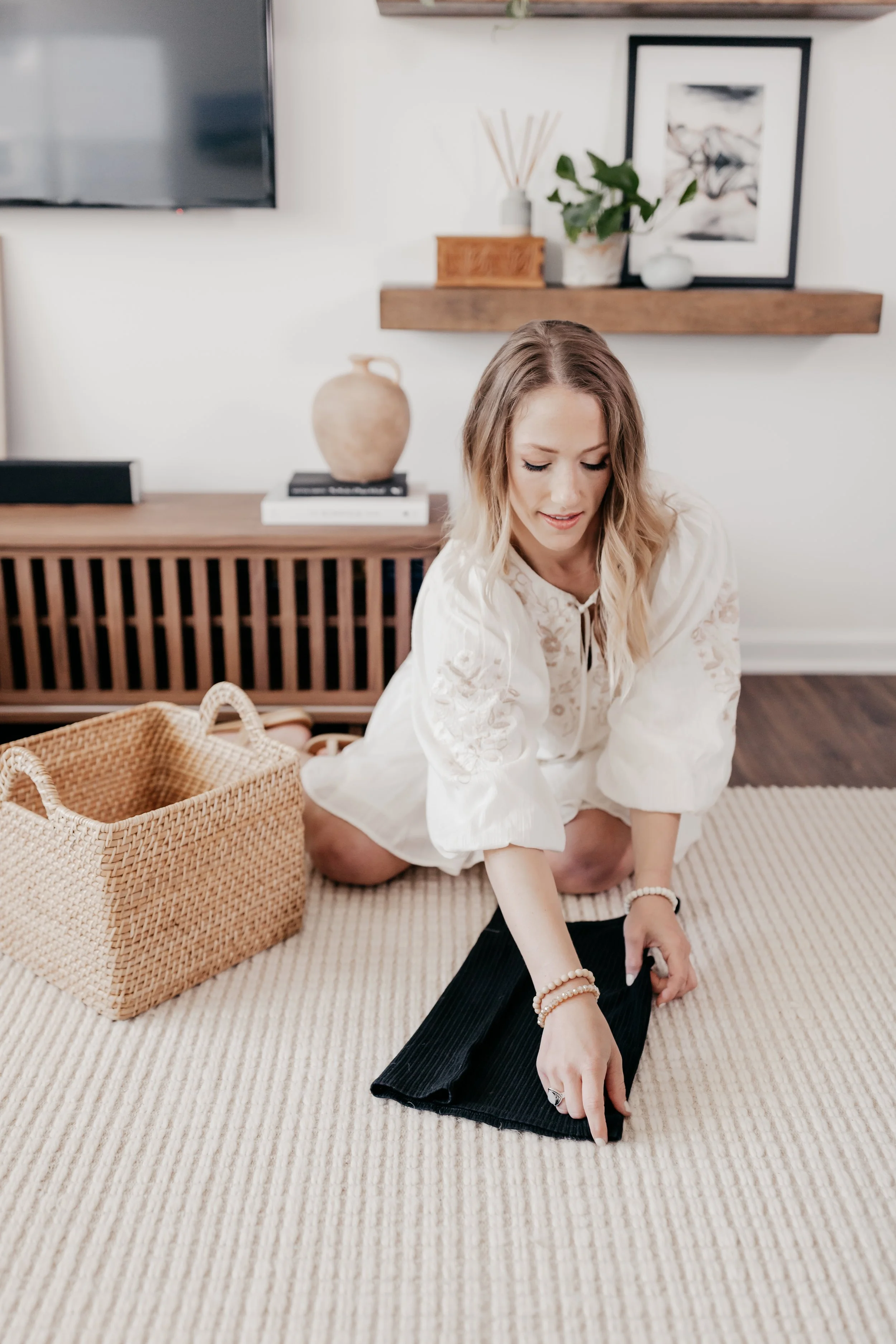 A woman is sitting on a beige rug in a living room, wearing a white lace top and black pants, with a wicker basket nearby and a decorative shelf with plants and art in the background.
