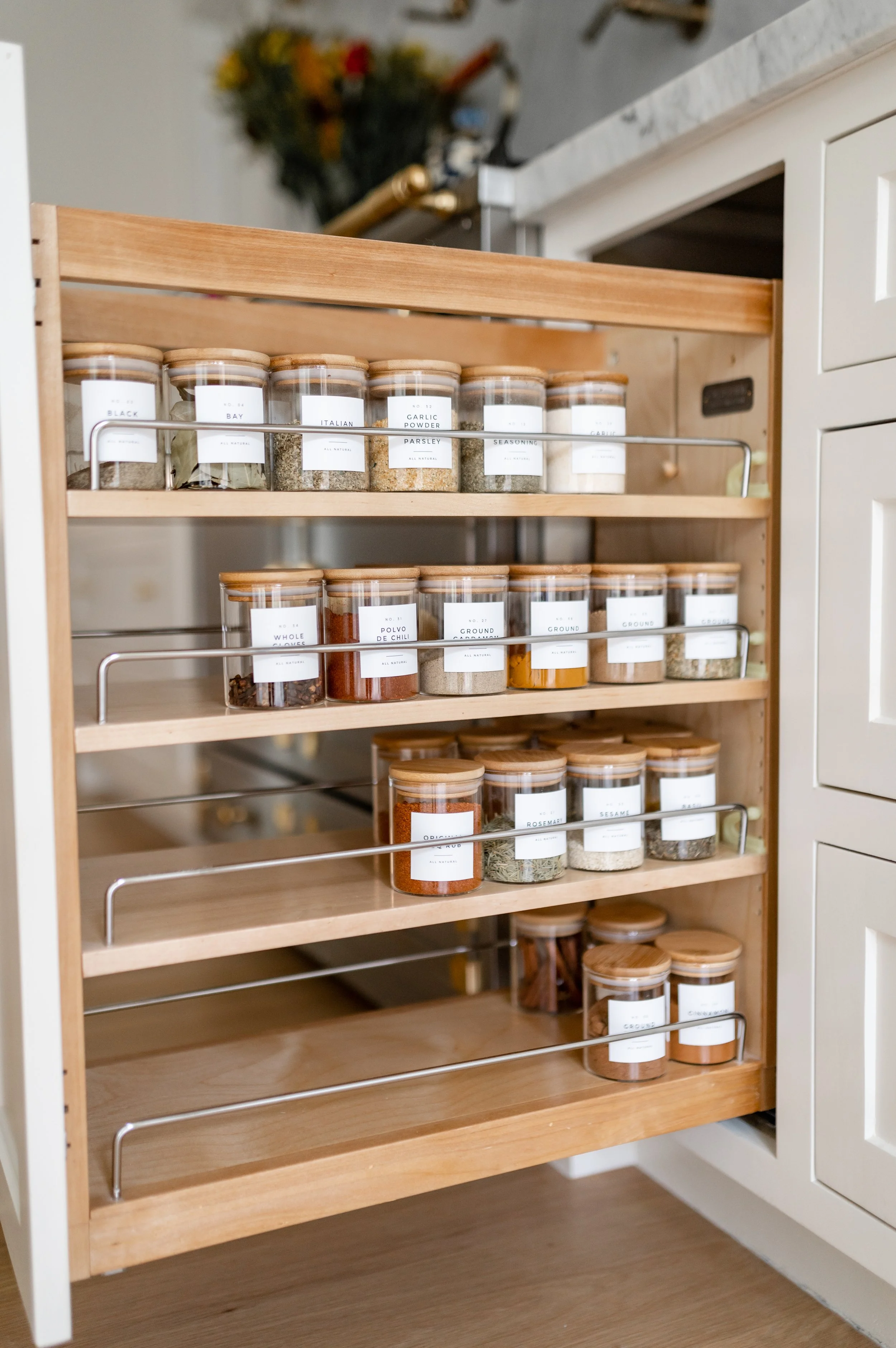 A wooden spice rack with glass jars filled with various spices and herbs in a kitchen. The jars have white labels with black text indicating the contents, such as black, bay, italian, garlic powder, parsley, seasoning, whole, polvo, de, chili, ground, rosemary, sesame, and more. The spice rack is positioned next to a white cabinet and a granite countertop.