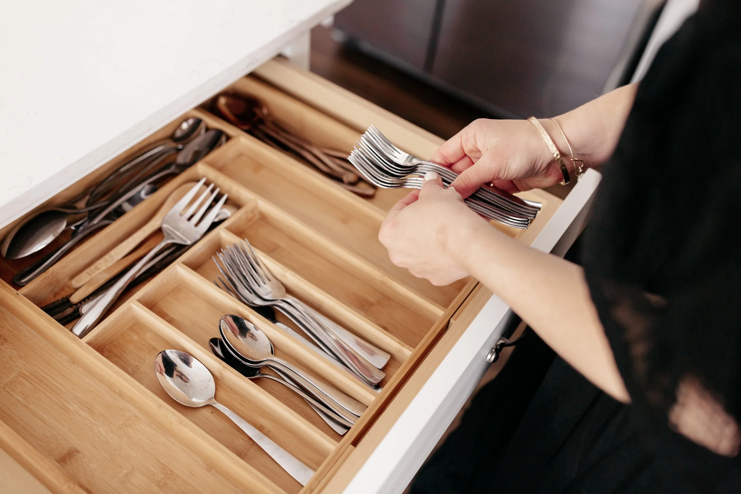 A person with a gold bracelet organizing silverware, including forks and spoons, in a wooden drawer with designated compartments.
