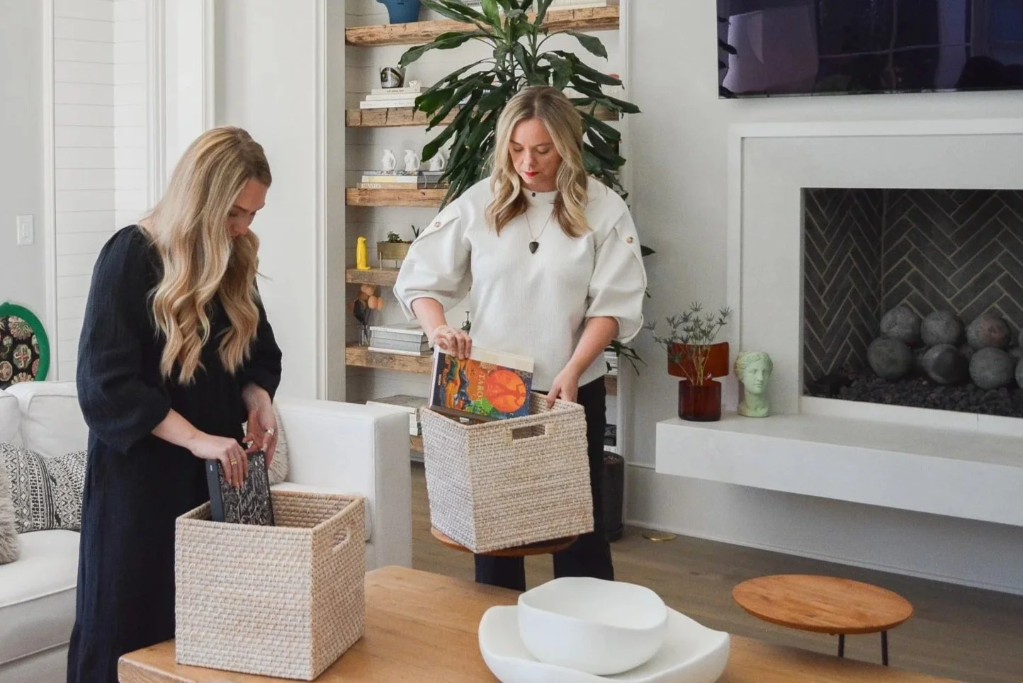 Two women organizing books and magazines into woven storage boxes on a wooden table inside a modern living room.