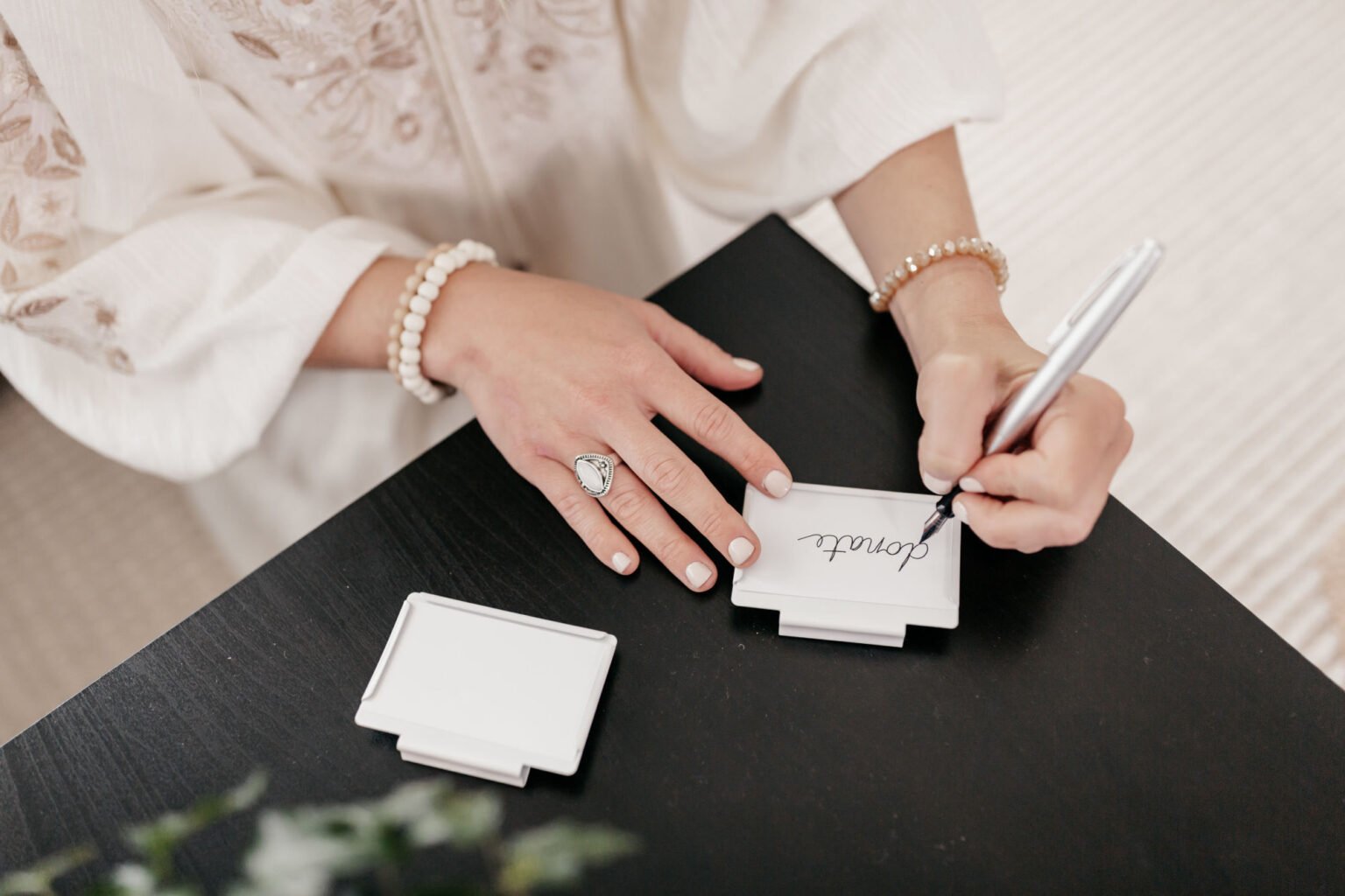 A woman writes the word 'love' on a small white card with a black marker. She wears a pearl bracelet, a large ring, and a beaded bracelet, and her sleeve is decorated with lace details. The card is placed on a black table with a stack of blank cards nearby.