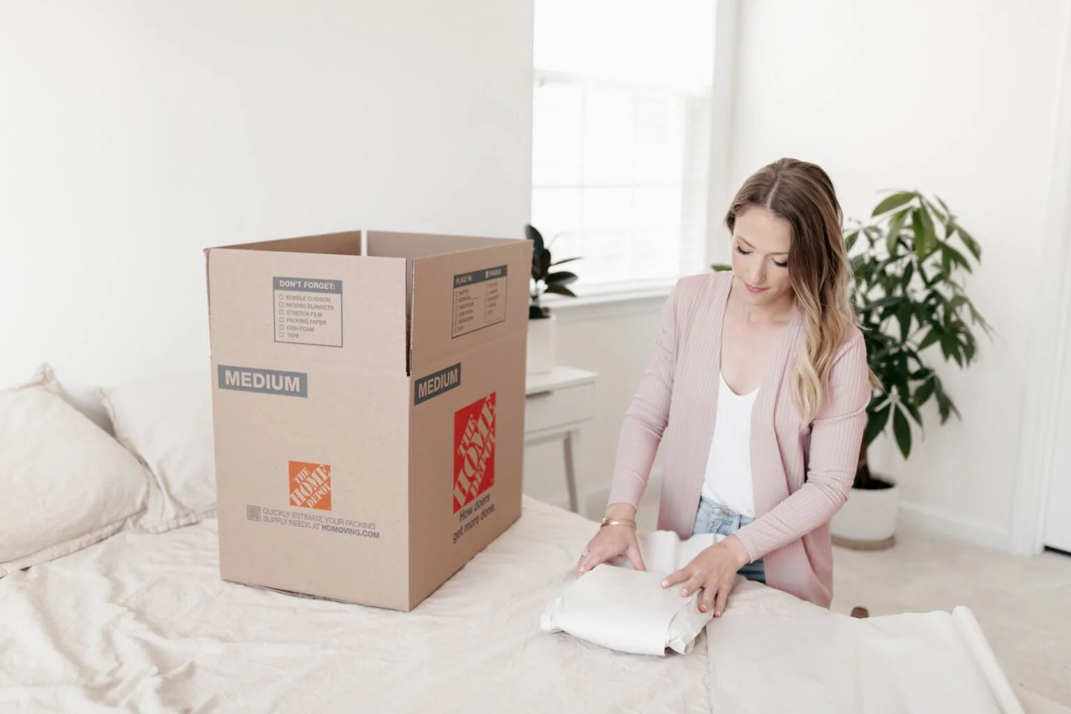 Woman packing items into a shipping box on a bed in a bright bedroom.
