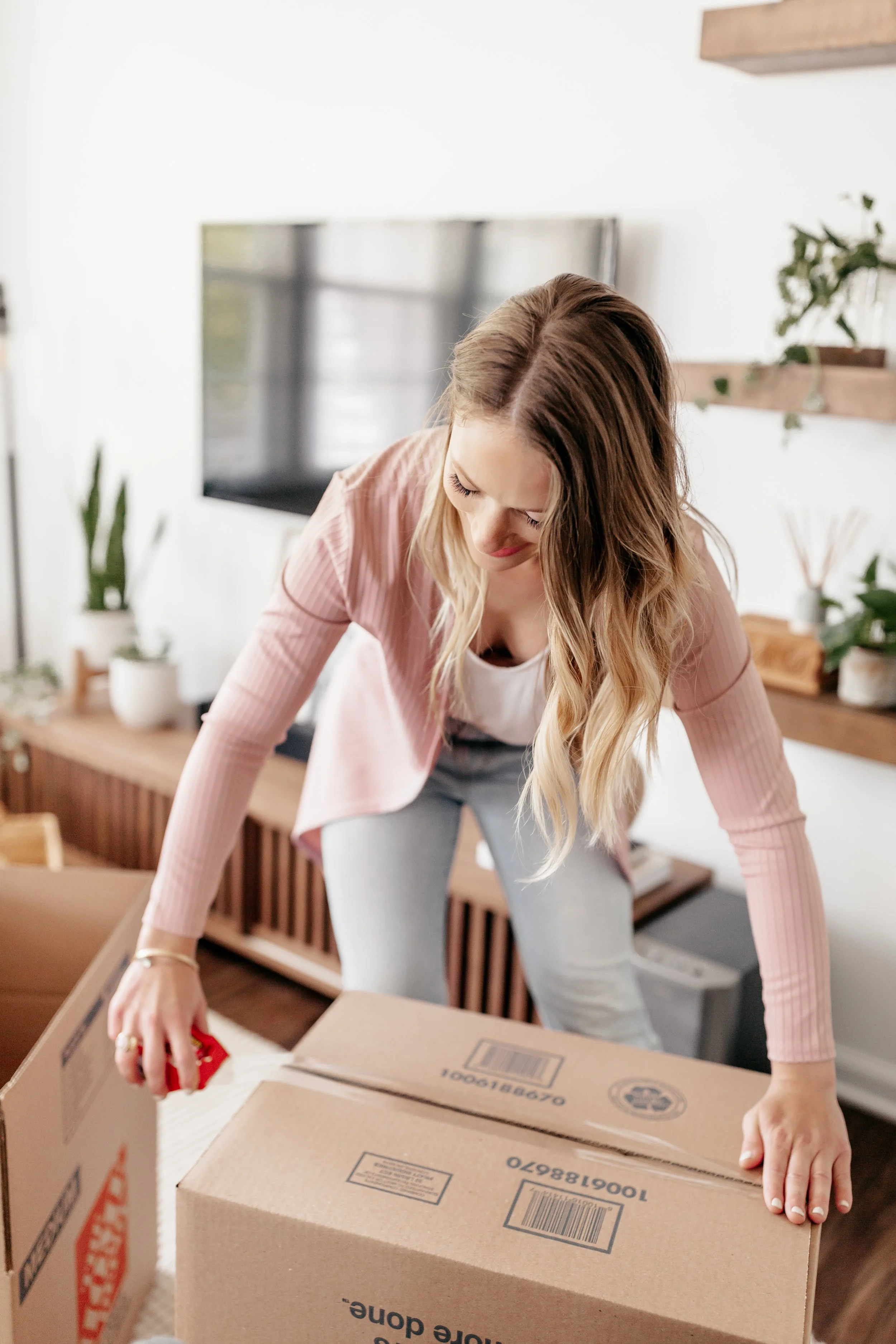 A young woman with long blonde hair, wearing a pink cardigan and gray pants, is unpacking moving boxes in a living room.