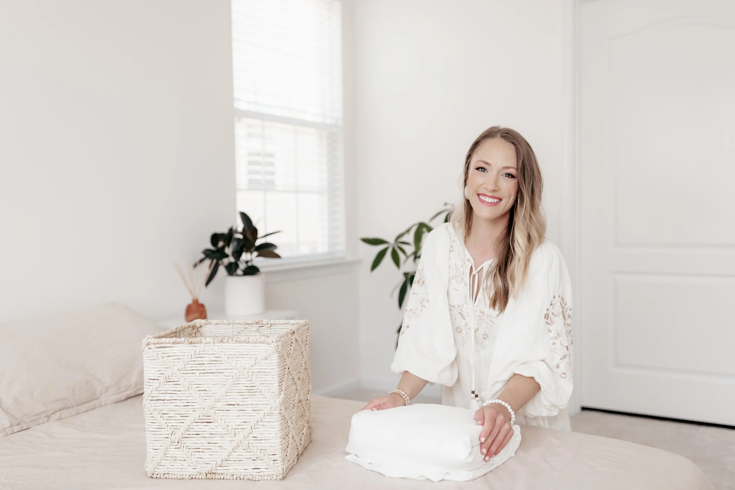 A woman with wavy blonde hair smiling while folding white linens in a bright, minimalist room with white walls and natural light.