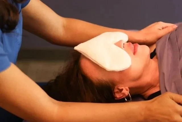 A woman lying down, wearing an eye mask, receiving care from a person in a blue shirt.