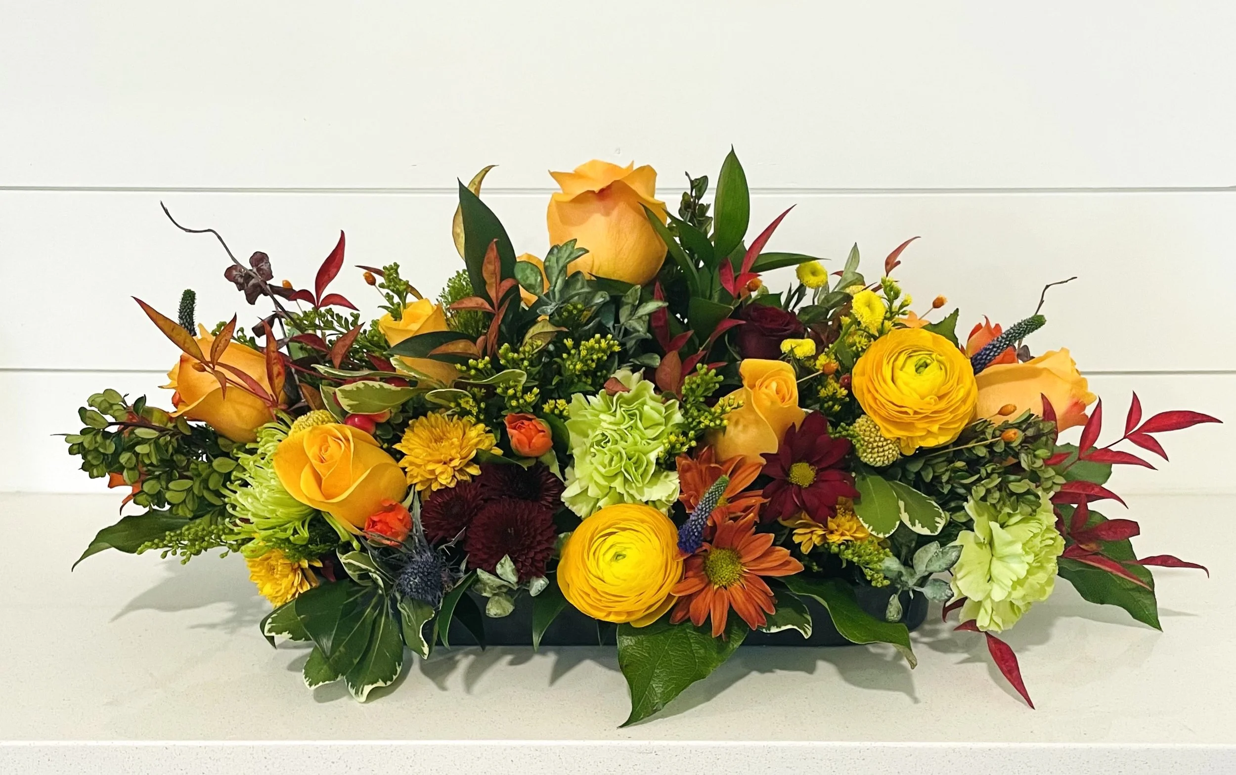 A colorful floral arrangement featuring yellow roses, orange and yellow daisies, dark maroon chrysanthemums, green and variegated foliage, and accents like blue thistle, set against a plain white background.