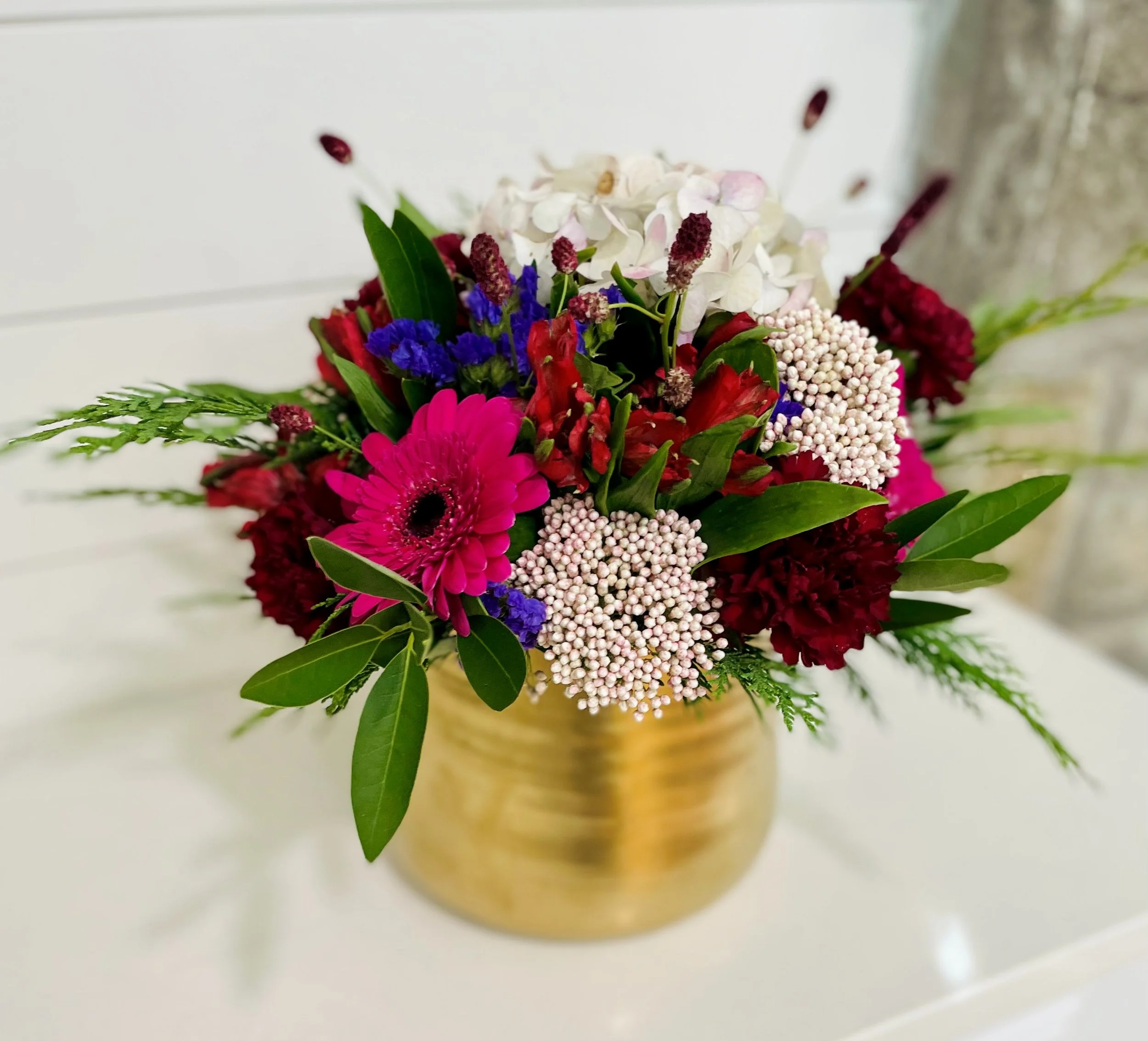 A colorful floral arrangement in a round gold vase, featuring pink gerbera daisies, white hydrangeas, red and purple flowers, and green foliage, placed on a white surface with a blurred background.
