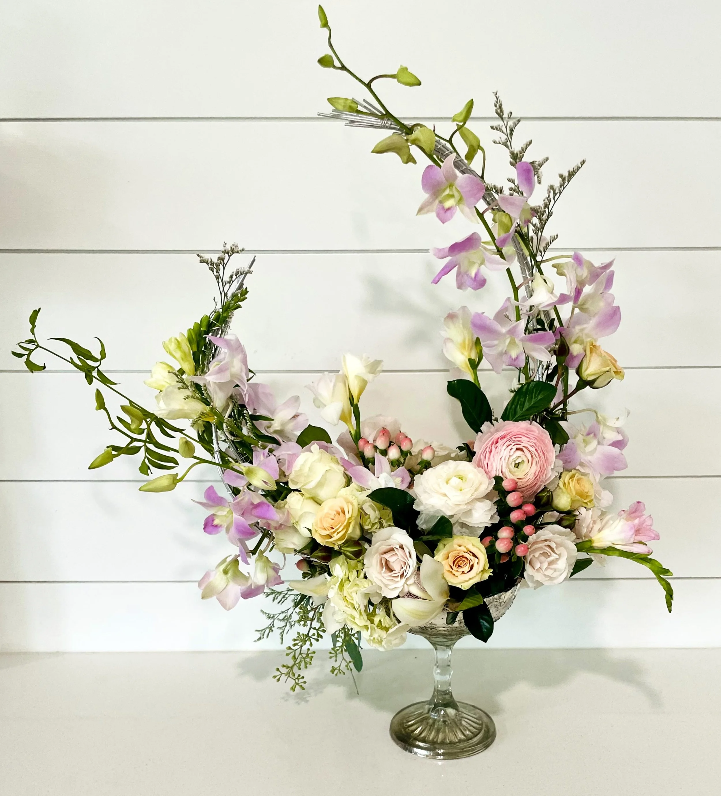 A floral arrangement in a glass vase featuring pink and white roses, pink ranunculus, and purple orchids with green foliage against a white wooden background.