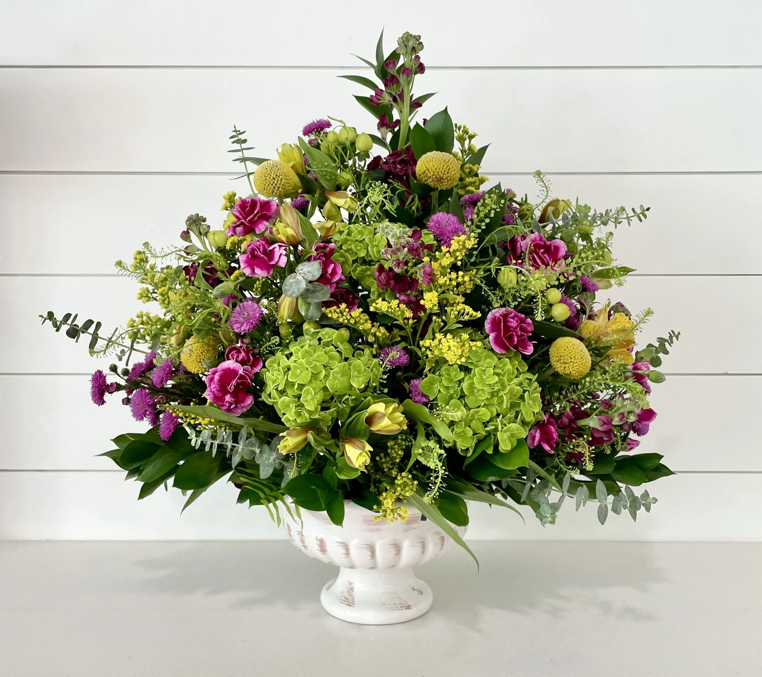 A large floral arrangement with pink, yellow, and green flowers in a white pedestal vase, against a white shiplap wall background.