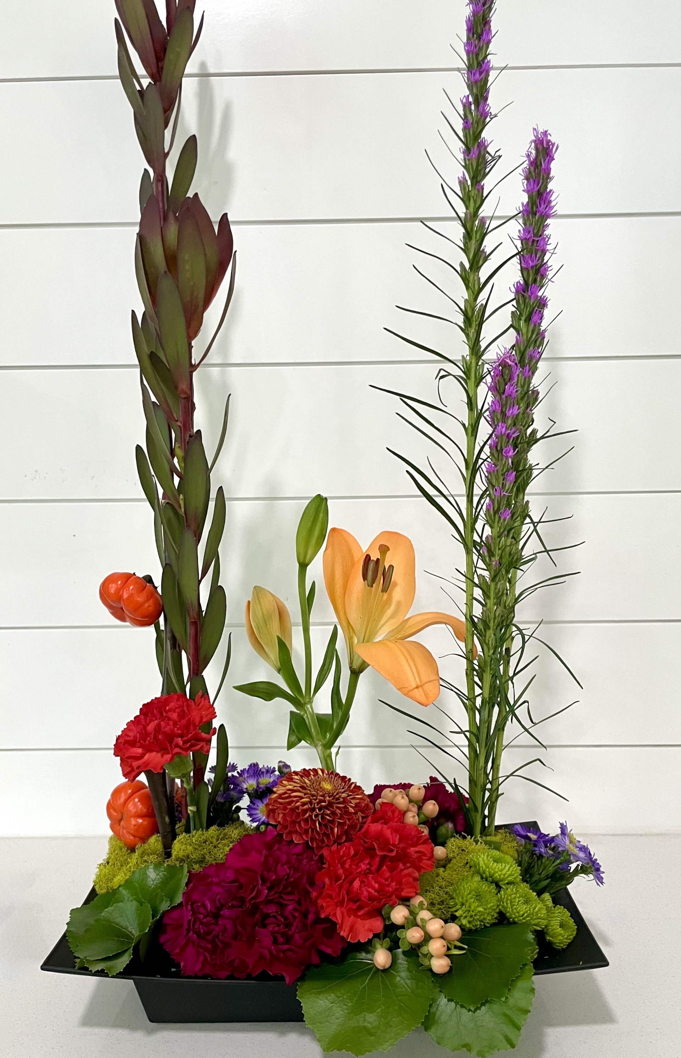 A floral arrangement with orange lilies, purple lavender, red carnations, green chrysanthemums, and berries, set in a black rectangular container against a white paneled background.